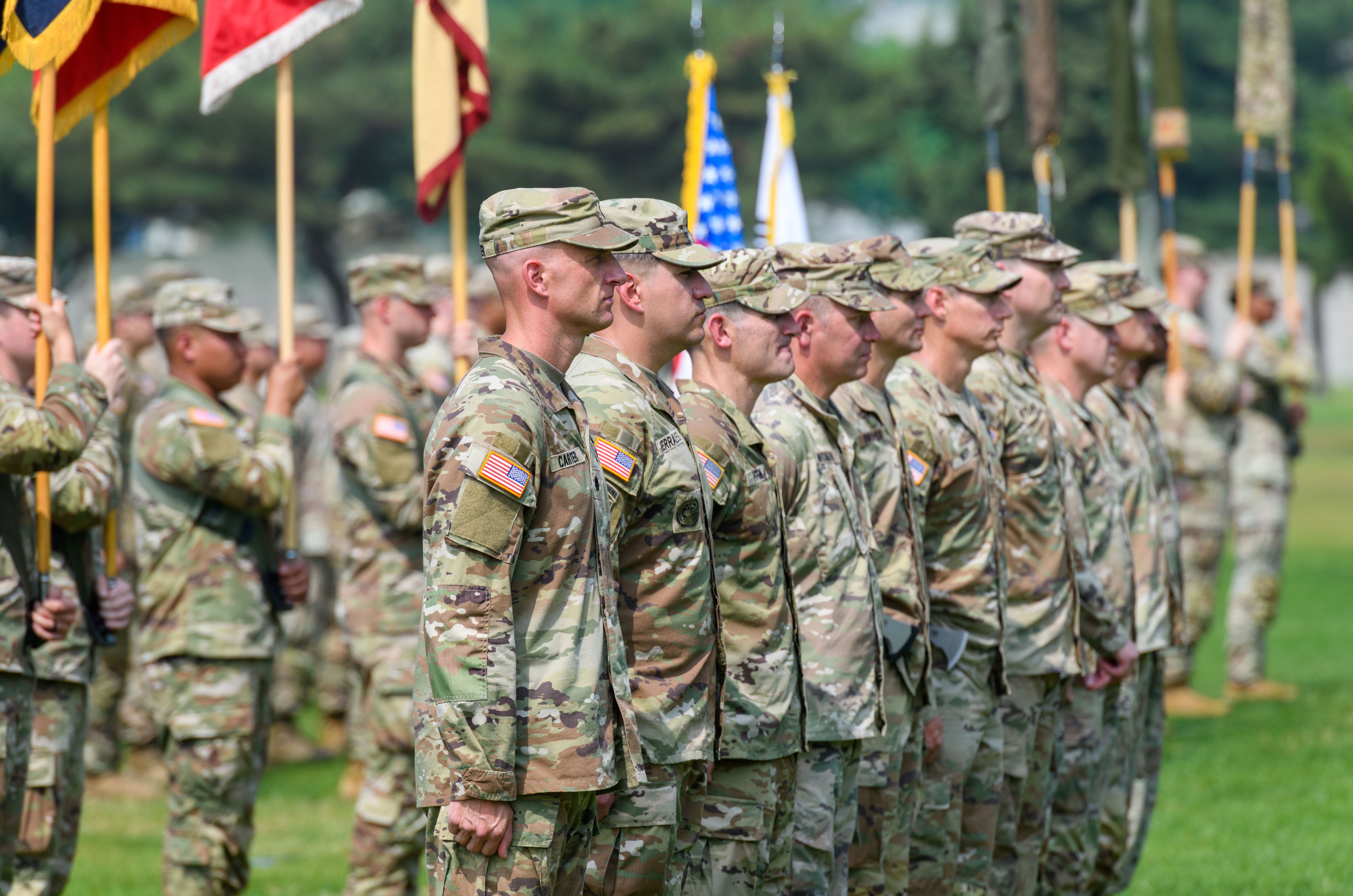 Soldiers in uniform stand in formation during a military ceremony, with flags visible in the background