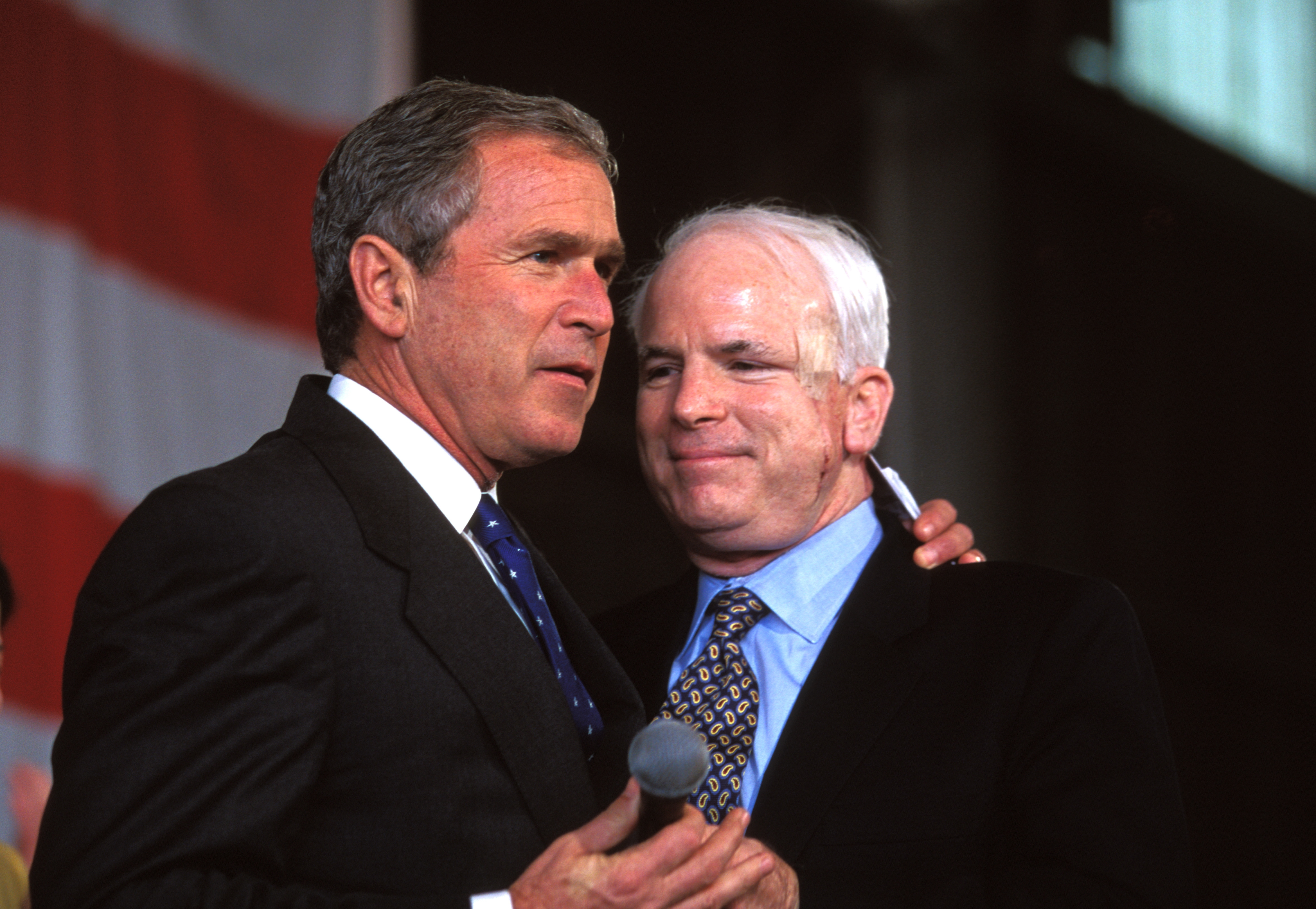 Two men in suits on stage, one holding a microphone, appear engaged in conversation at a public event