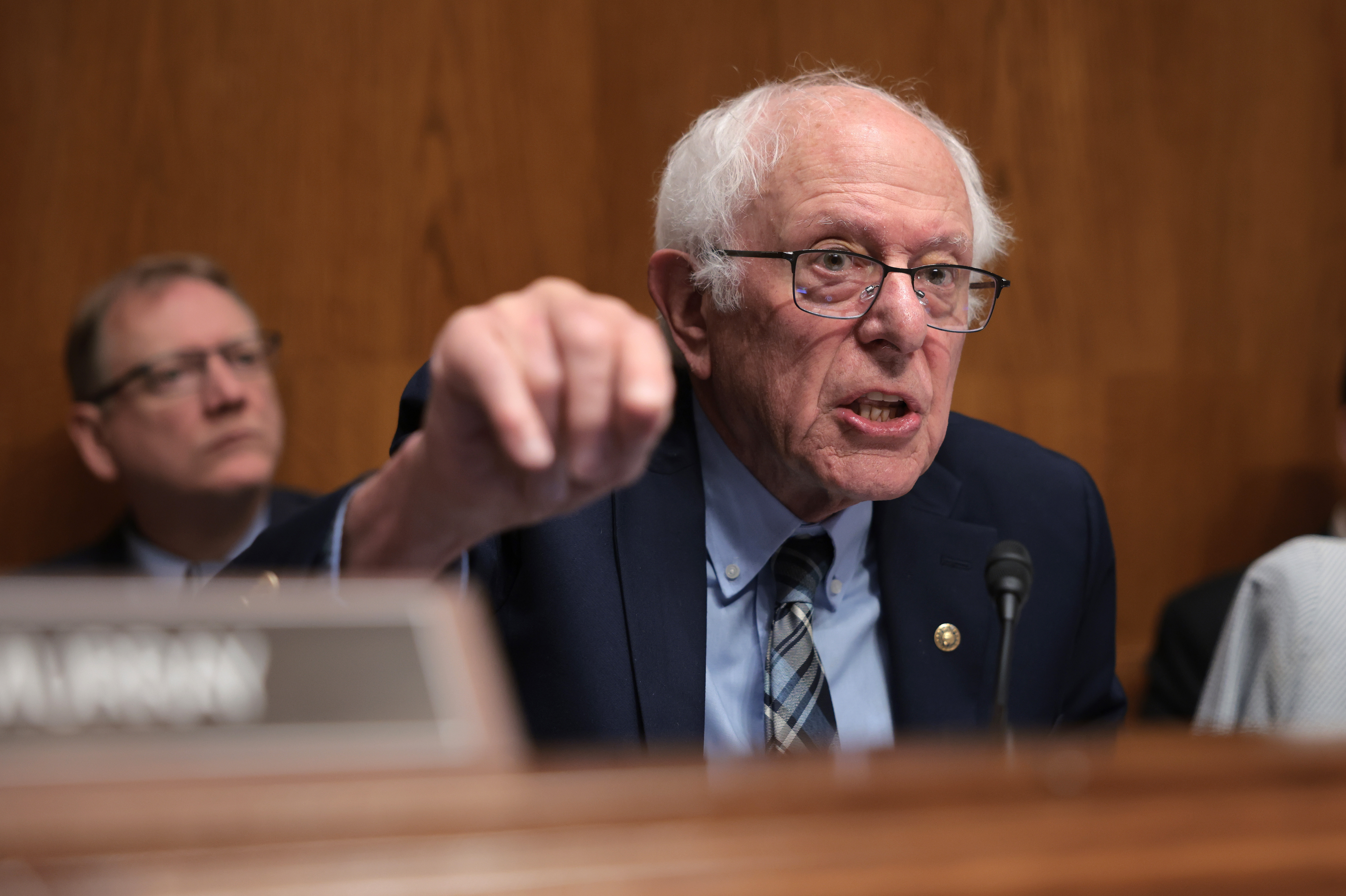 An older man in a suit gestures emphatically while speaking at a hearing, with people seated behind him