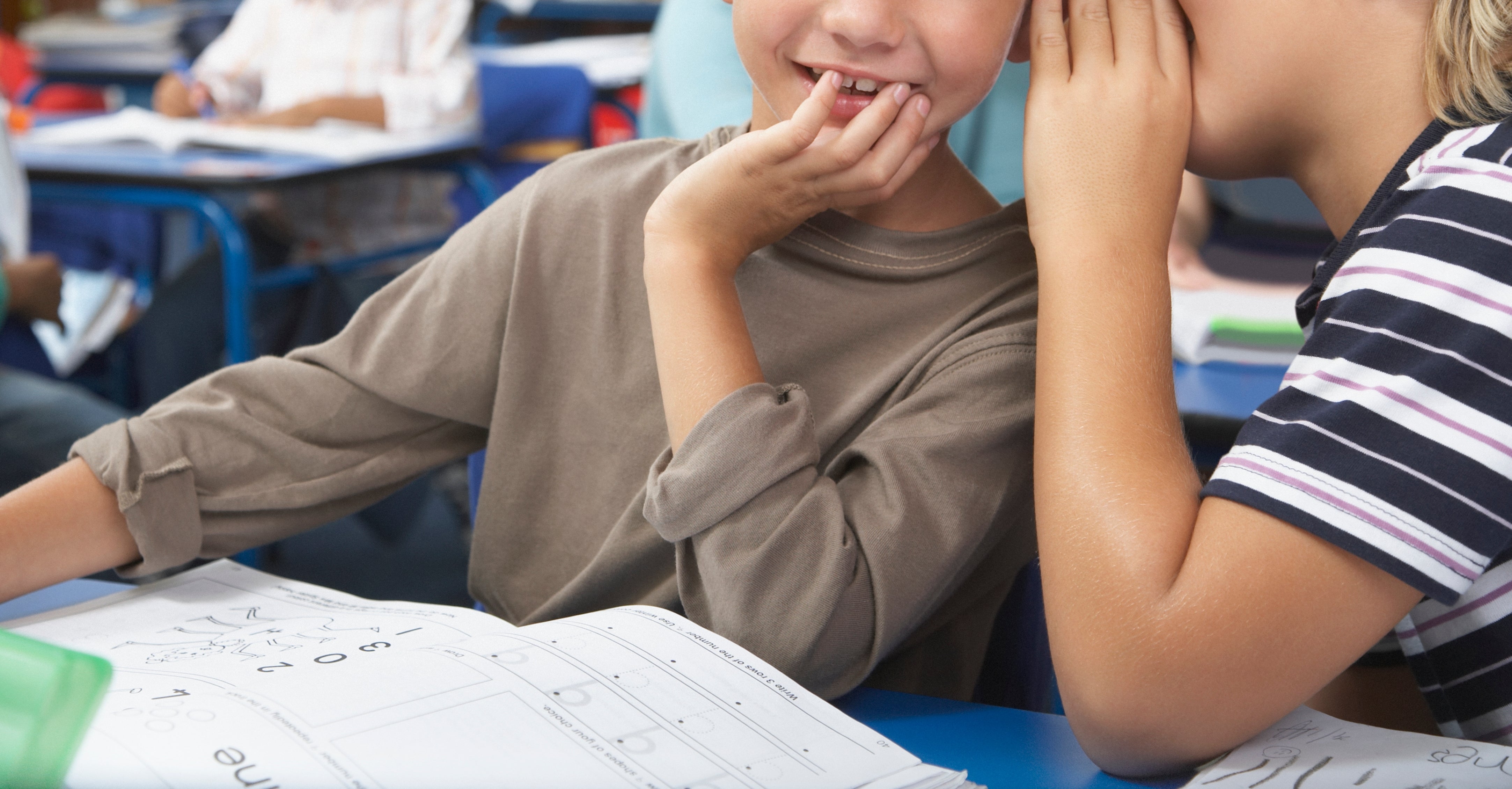Two young boys are in a classroom; one boy is whispering to the other, who is smiling. Other students are focused on their work at desks