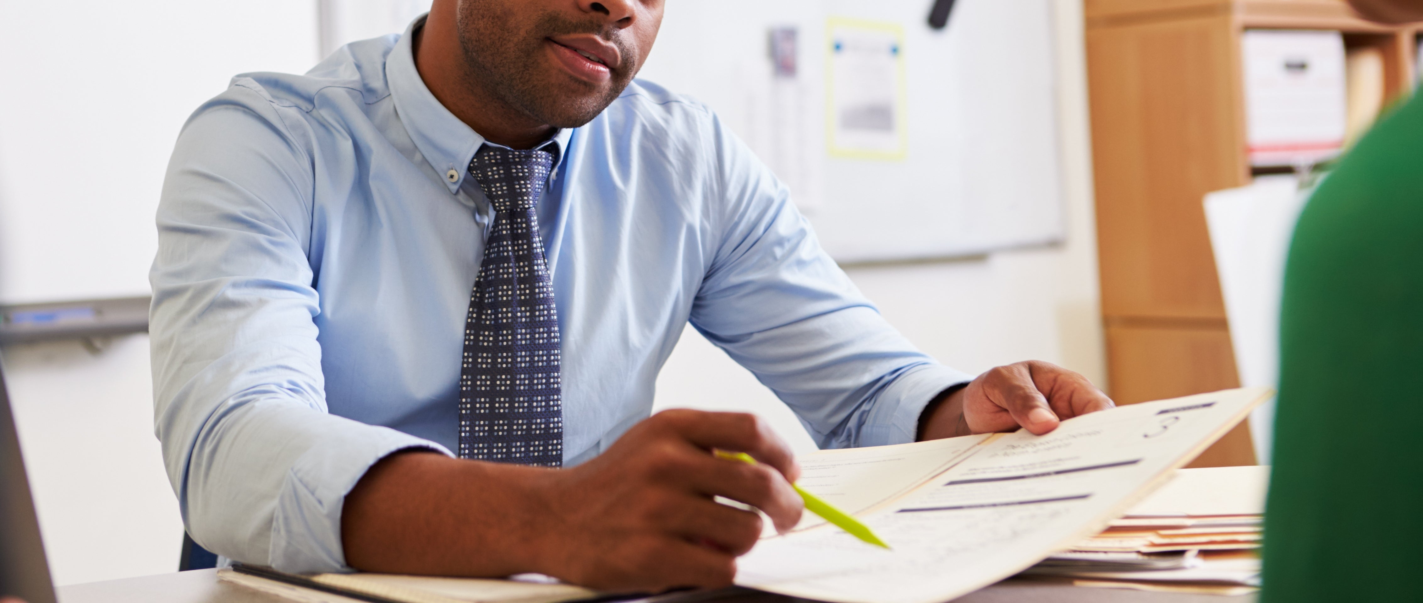 A person in a shirt and tie sits at a desk with papers, attentively speaking to two individuals across from them. A whiteboard is in the background