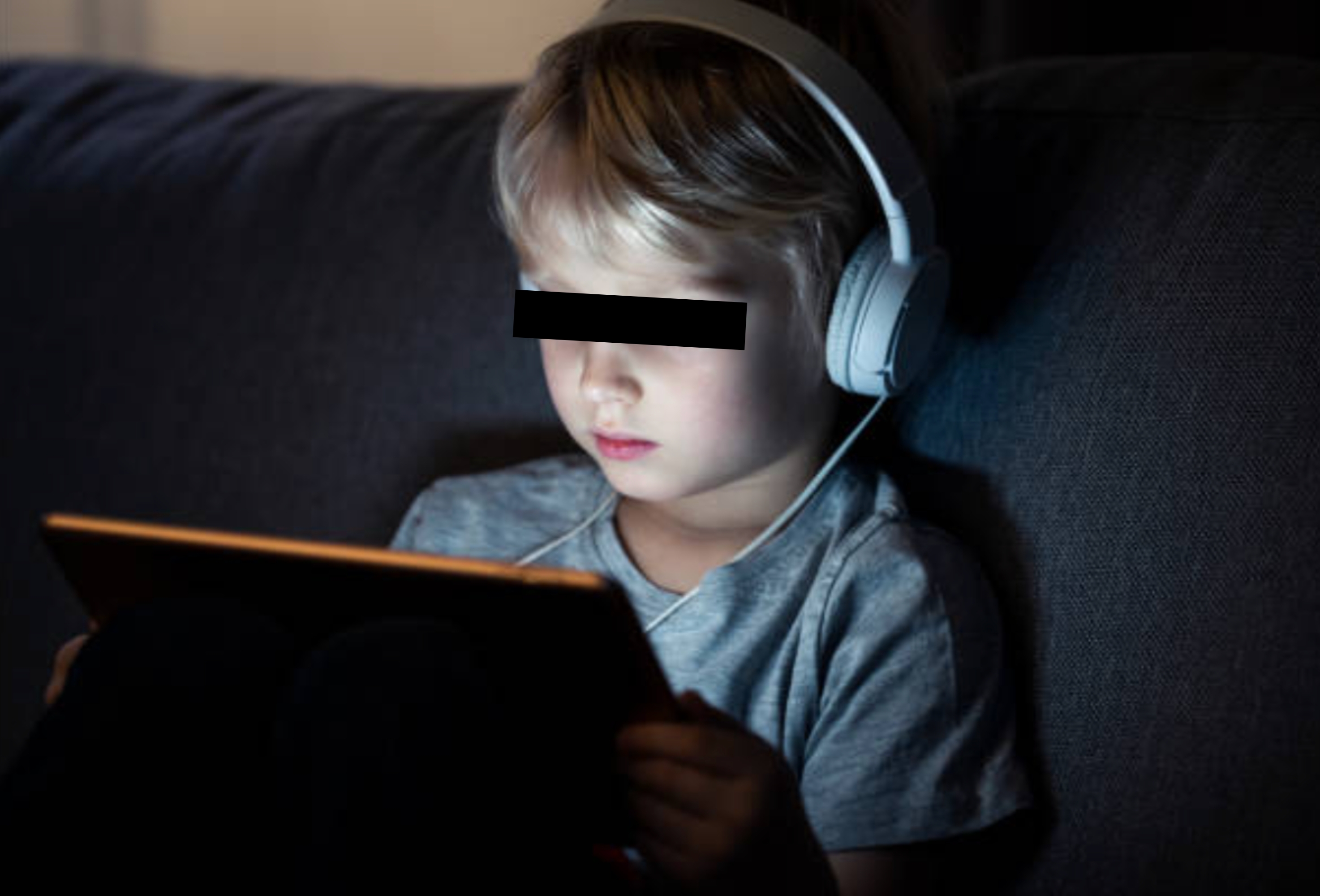 Young child wearing headphones, focused on a tablet screen while sitting on a couch, showing concentration and engagement with the device