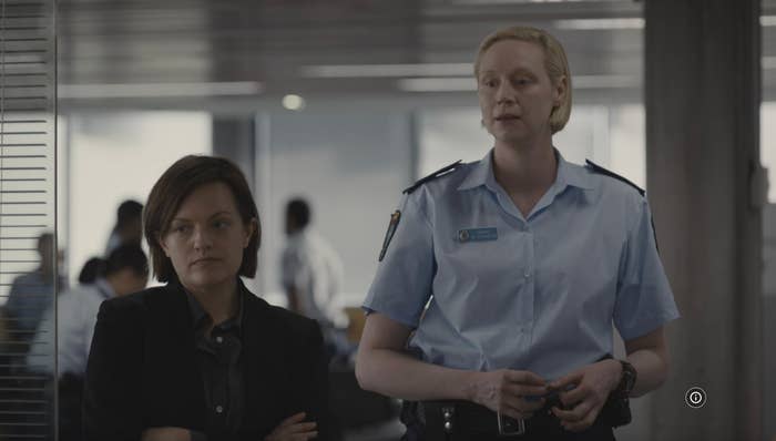 Two people are standing indoors, one in a dark suit and the other in a police uniform, engaged in conversation