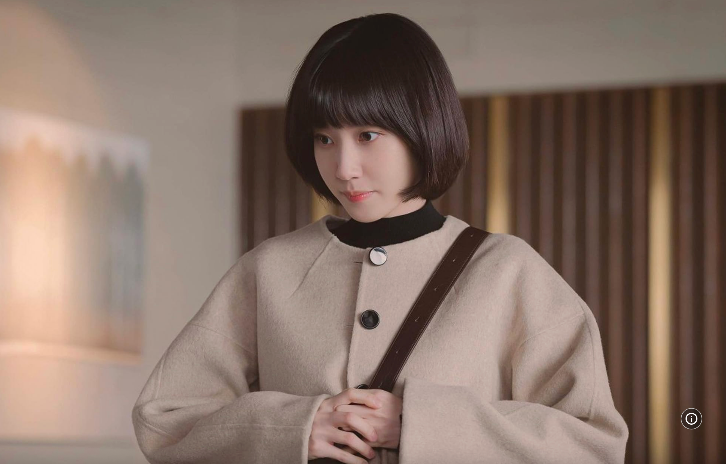 Person with a bob haircut and bangs, wearing a chic, buttoned-overcoat and crossbody bag, indoors