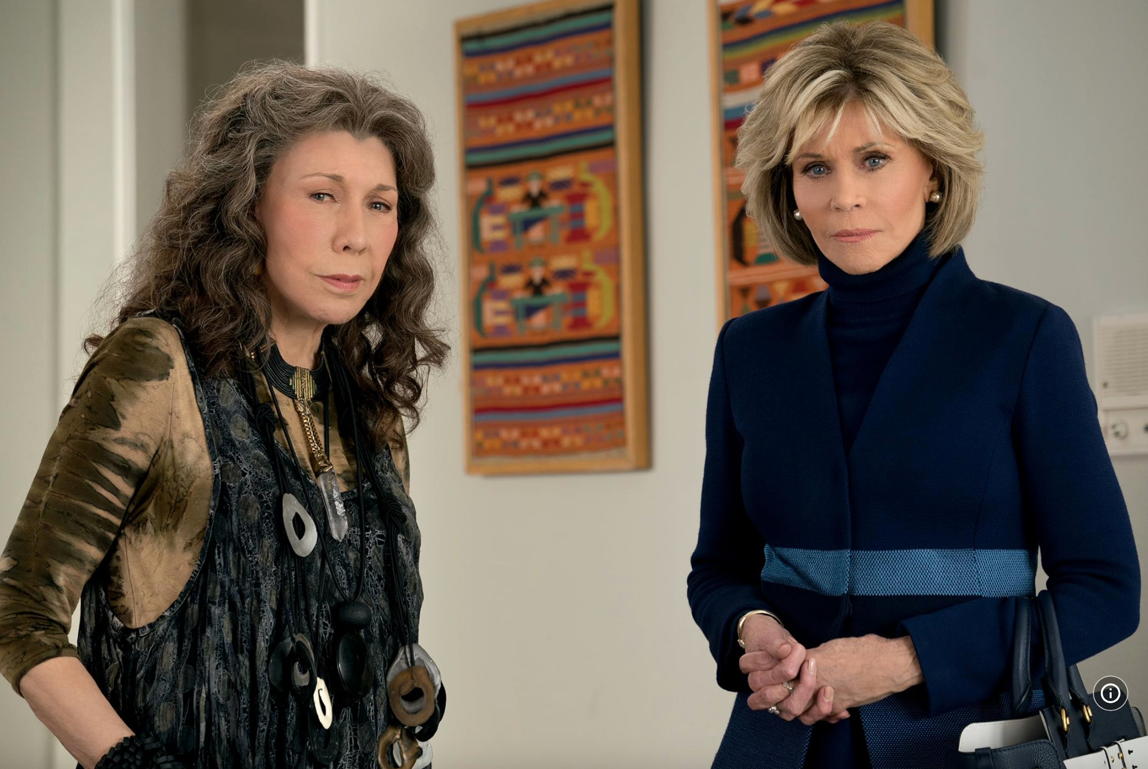 Two women in a stylish, modern setting. One wears layered clothing with bold necklaces; the other wears a fitted outfit with a belt