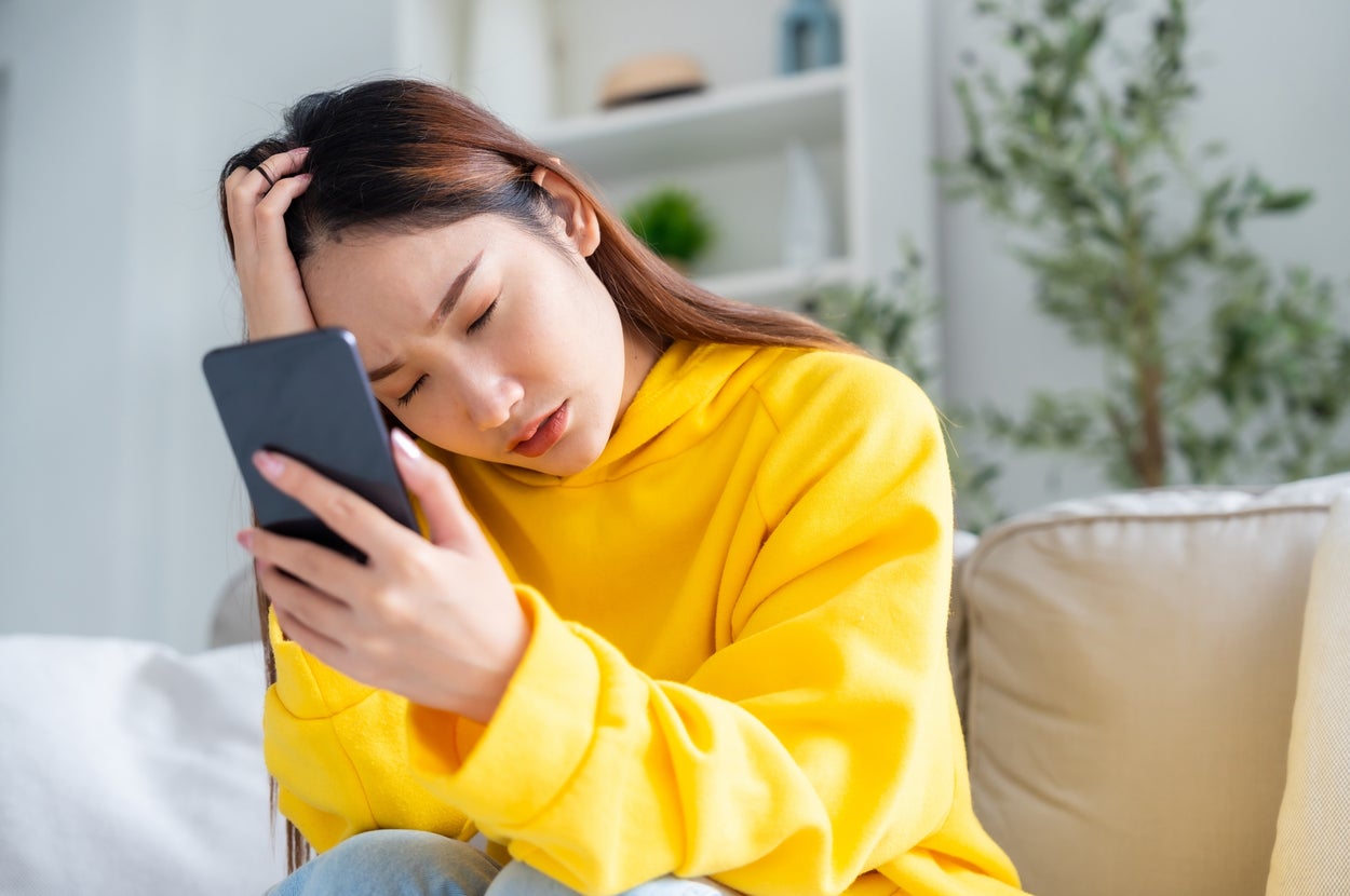 女性がスマートフォンを見ながら悩んでいる様子。部屋のソファに座り、リラックスした服装をしています。