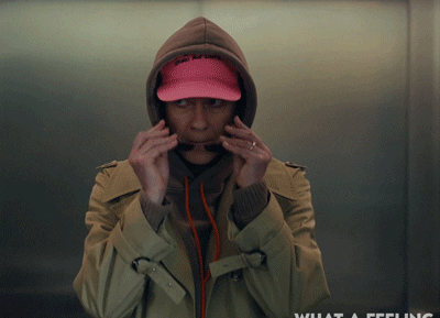 Person in layered casual clothes and pink cap dances energetically in an elevator. The words "What a feeling" are displayed in the lower right corner