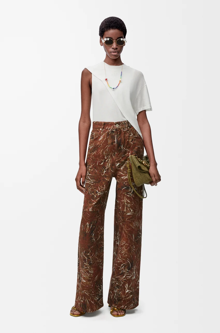 Person in asymmetrical white top, patterned wide-leg pants, sunglasses, necklace, and holding a textured clutch on a plain background