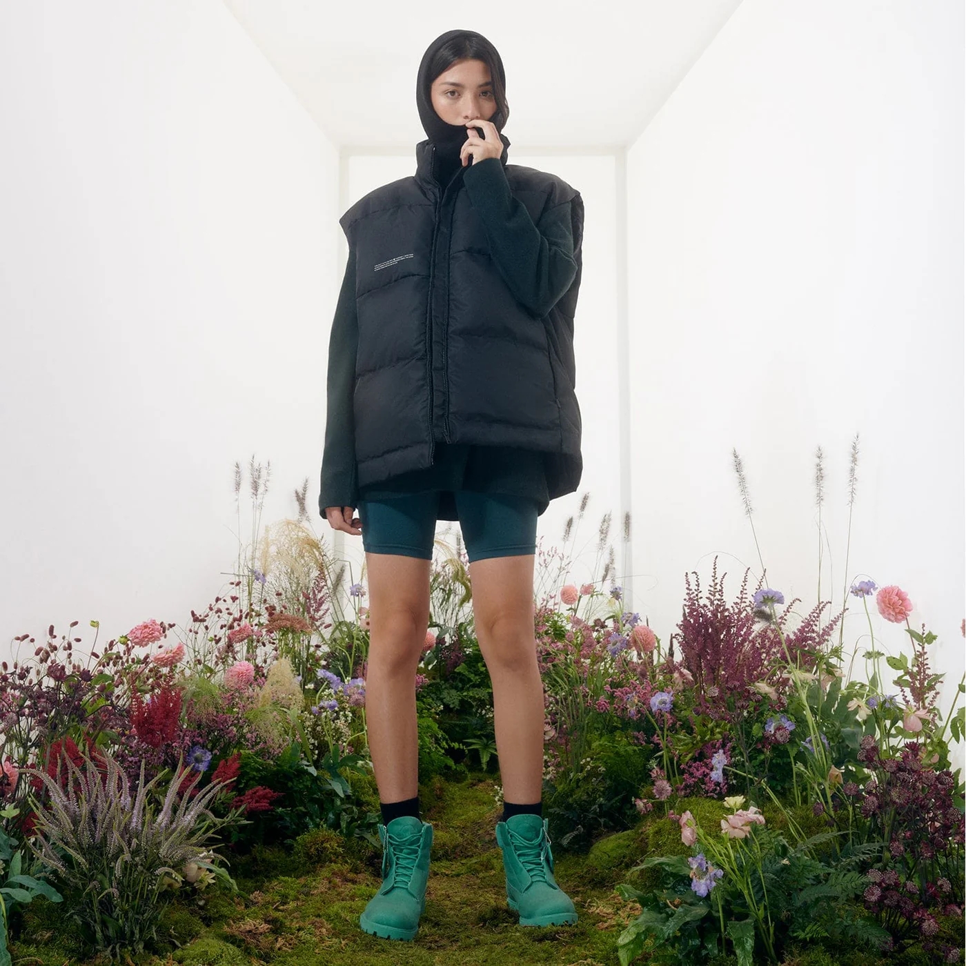 Person in puffy jacket and shorts stands on indoor garden path, surrounded by flowers, holding a phone