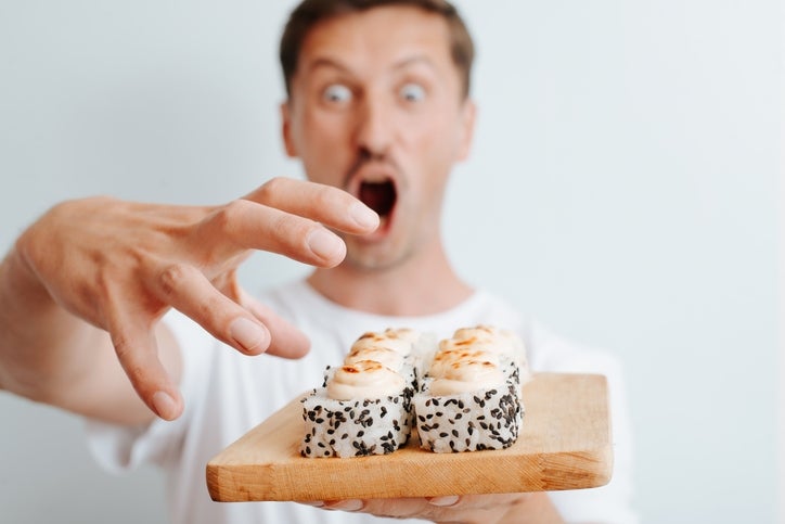 Seorang pria meraih gulungan sushi dengan penuh semangat di papan kayu, tampak terkejut