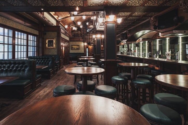 Interior pub yang luas dengan meja kayu bundar, bangku empuk, dan bar panjang dengan keran. Suasana remang-remang menunjukkan suasana nyaman dan ramah
