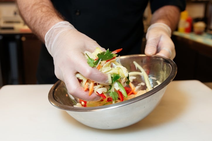 Orang yang memakai sarung tangan mencampur salad segar dengan bumbu dan sayuran dalam mangkuk logam