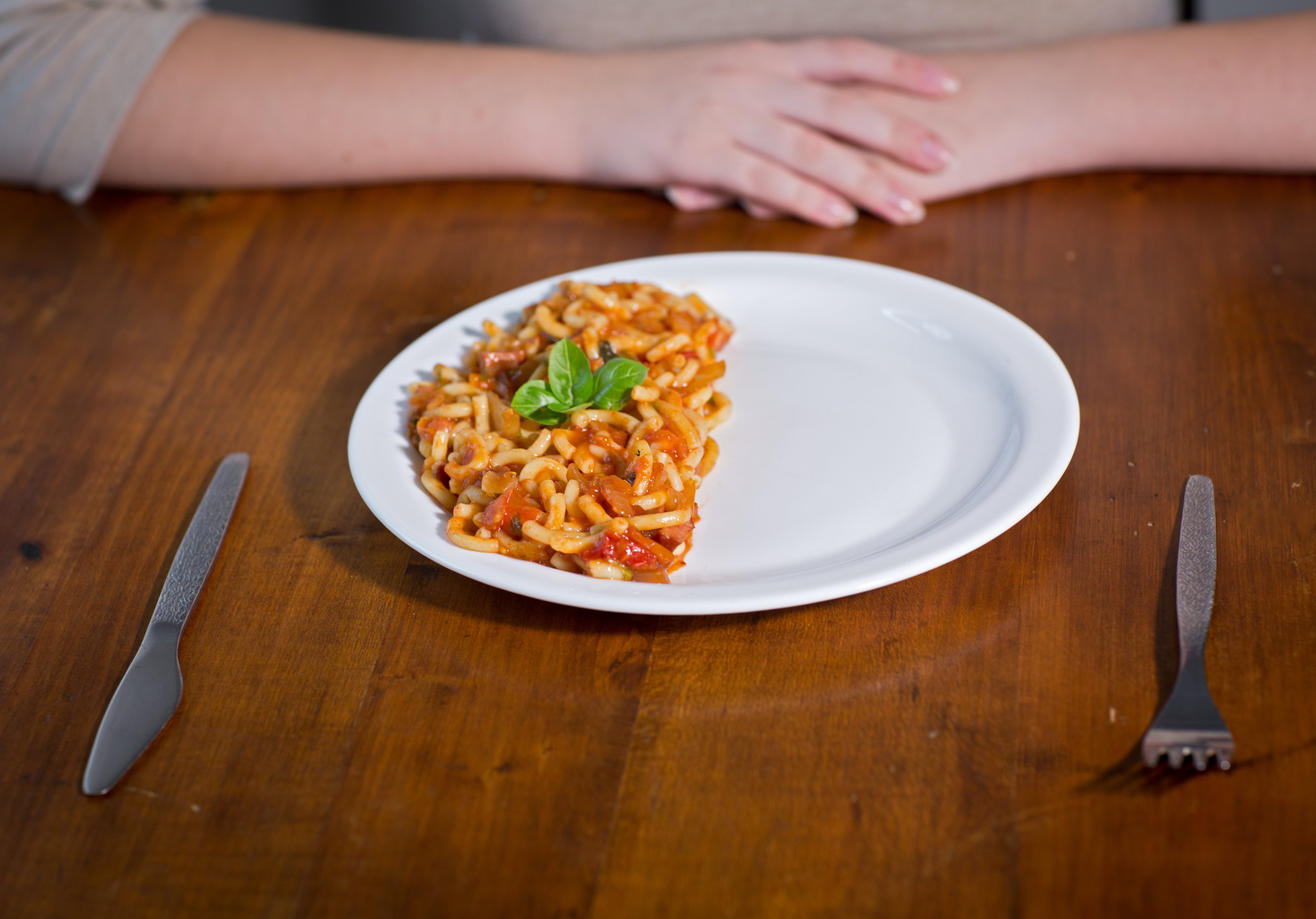 Piring dengan setengah porsi pasta yang dihias dengan kemangi di atas meja kayu; tangan seseorang di latar belakang, pisau dan garpu di samping piring