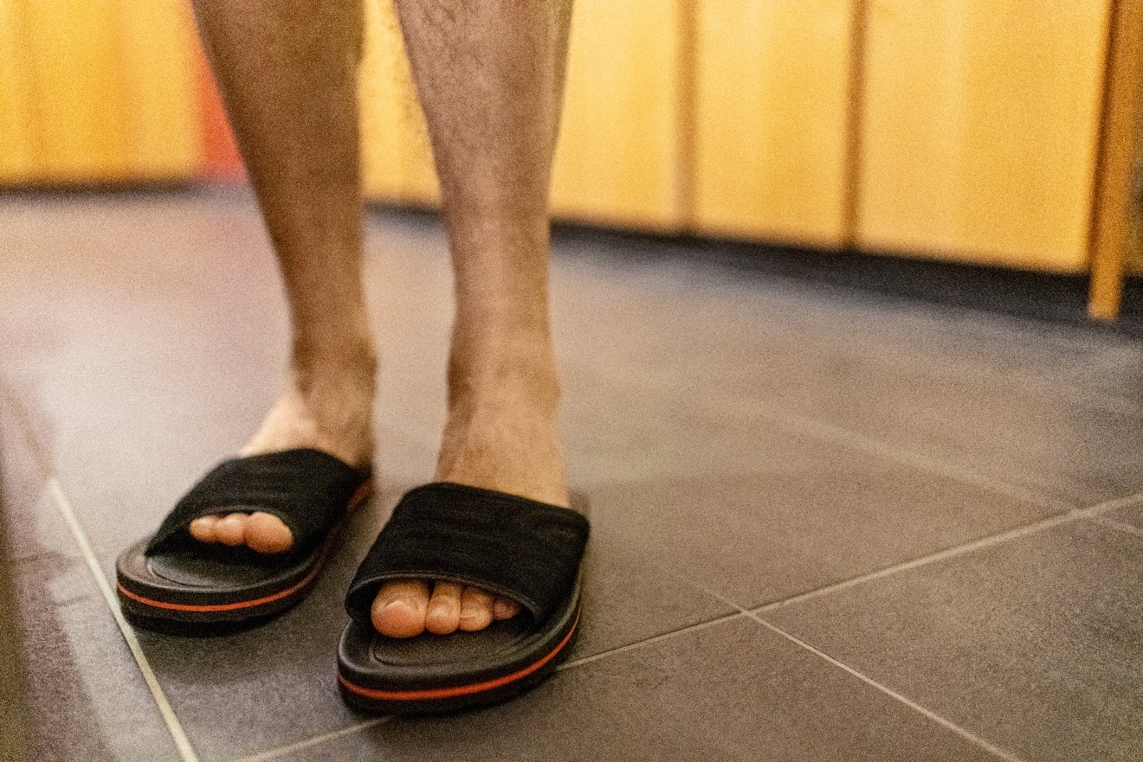 Orang yang berdiri di dalam ruangan mengenakan sandal geser hitam, dilihat dari mata kaki ke bawah. Lantai ubin dan latar belakang kayu terlihat
