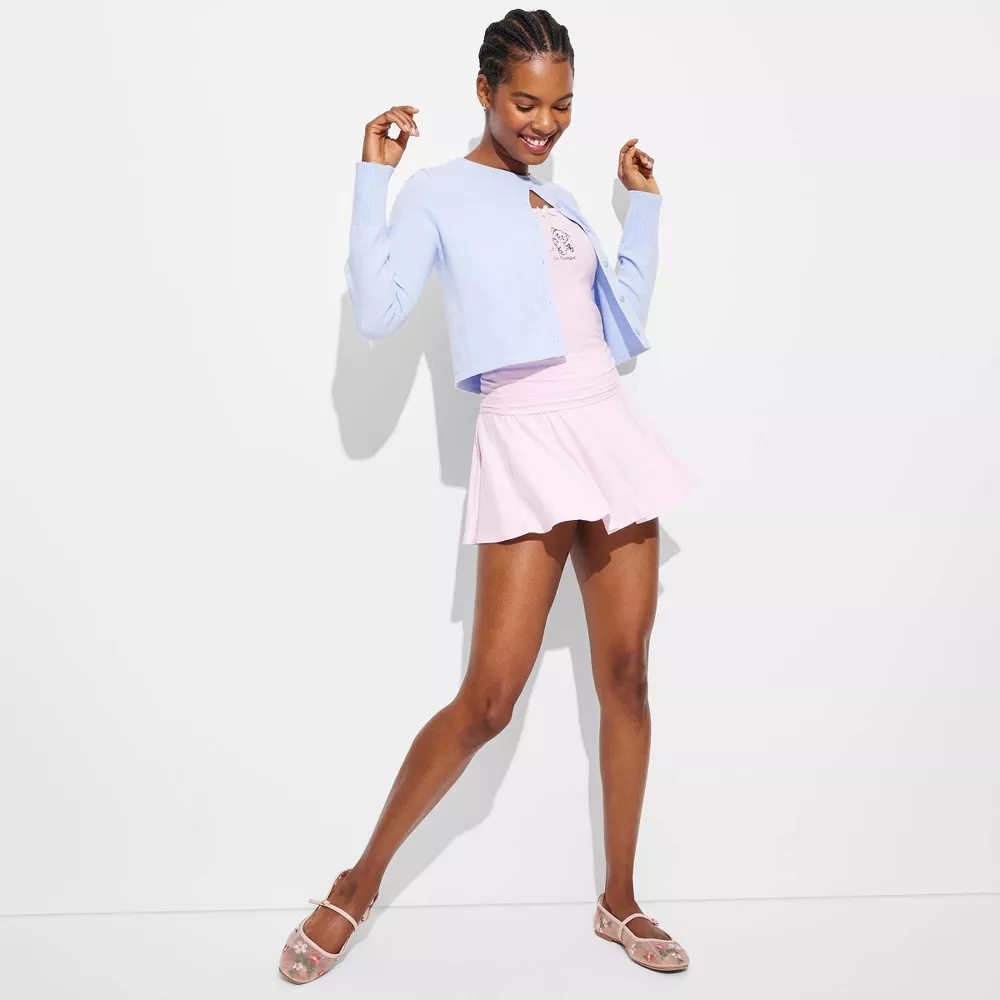 Person in a stylish, light cropped sweater and short skirt, posing playfully. They are wearing elegant, strappy sandals