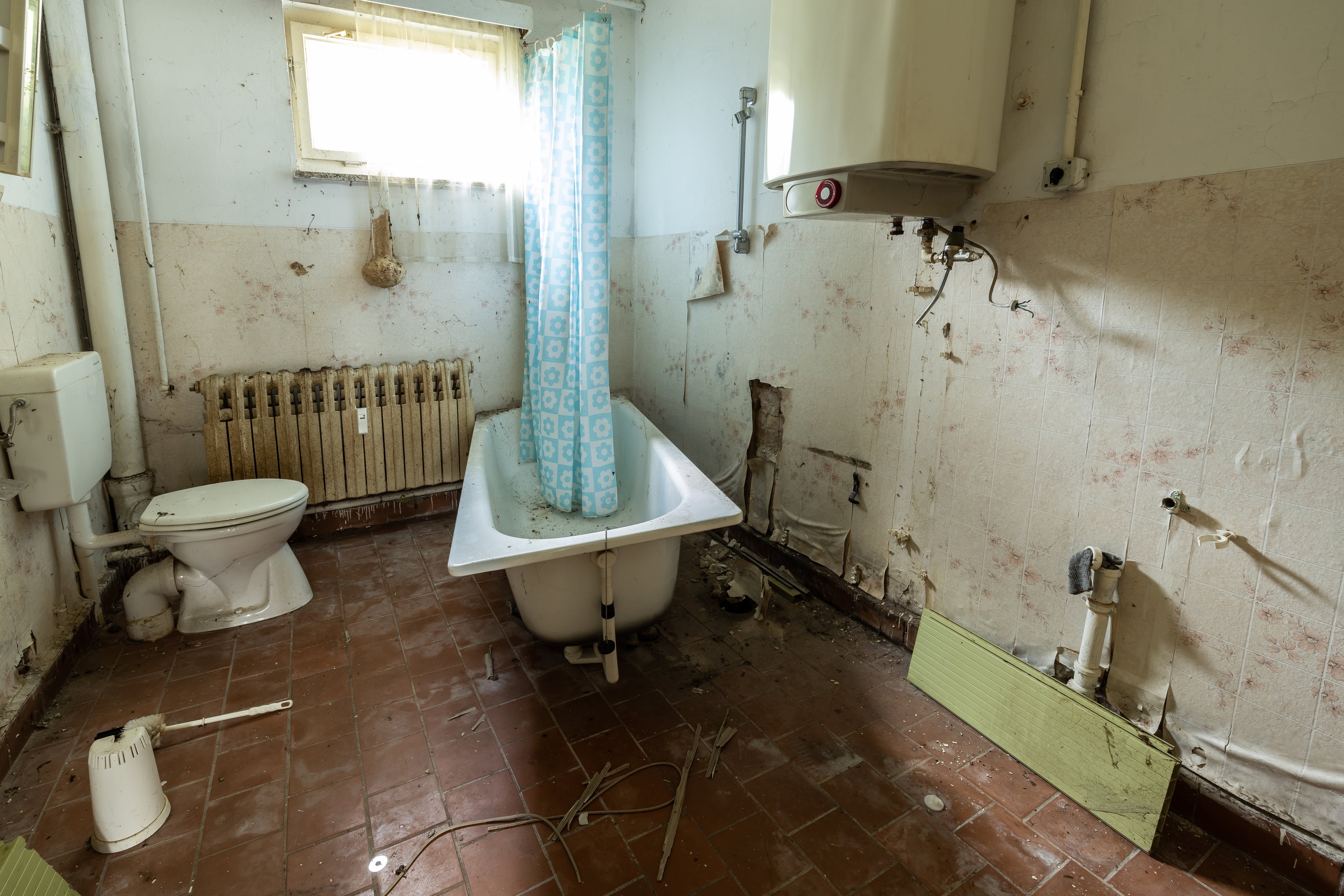 Kamar mandi tua dan kumuh dengan pipa terbuka, ubin pecah, bak mandi bertirai, dan toilet bekas, menandakan kelalaian dan kerusakan