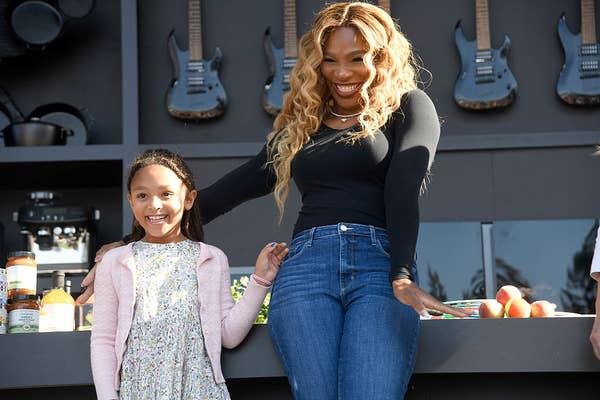 A smiling woman in a casual outfit stands with a young girl, holding hands. Guitars hang in the background
