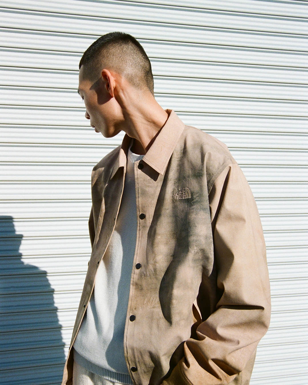 Person facing sideways wearing a casual jacket over a sweater, standing in front of a corrugated metal wall