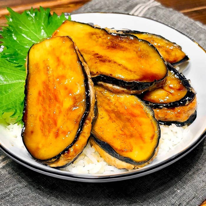 お皿に盛られた焼きナスの照焼きが白ご飯の上に乗っている料理。隣に緑の葉が添えられています。
