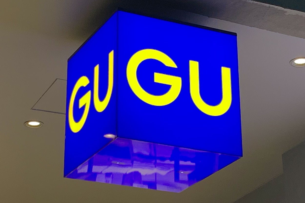 Illuminated cube sign with yellow "GU" letters on a dark background, seen hanging from a ceiling indoors