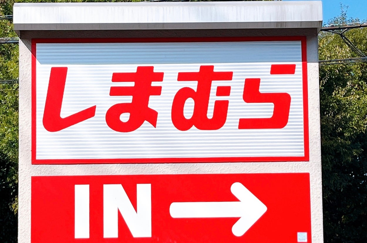 しまむら駐車場の入り口を示す看板。白地に赤色で「しまむら」「IN →」と書かれている。背景は緑の木々。