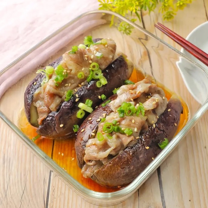 ガラス皿に盛られた焼きナスの上に薄切り肉とネギがトッピングされた料理。テーブルに箸も置かれている。