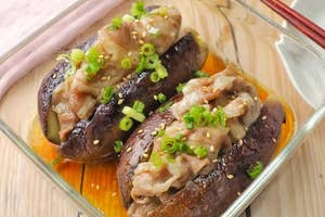 ガラス皿に盛られた焼きナスの上に薄切り肉とネギがトッピングされた料理。テーブルに箸も置かれている。