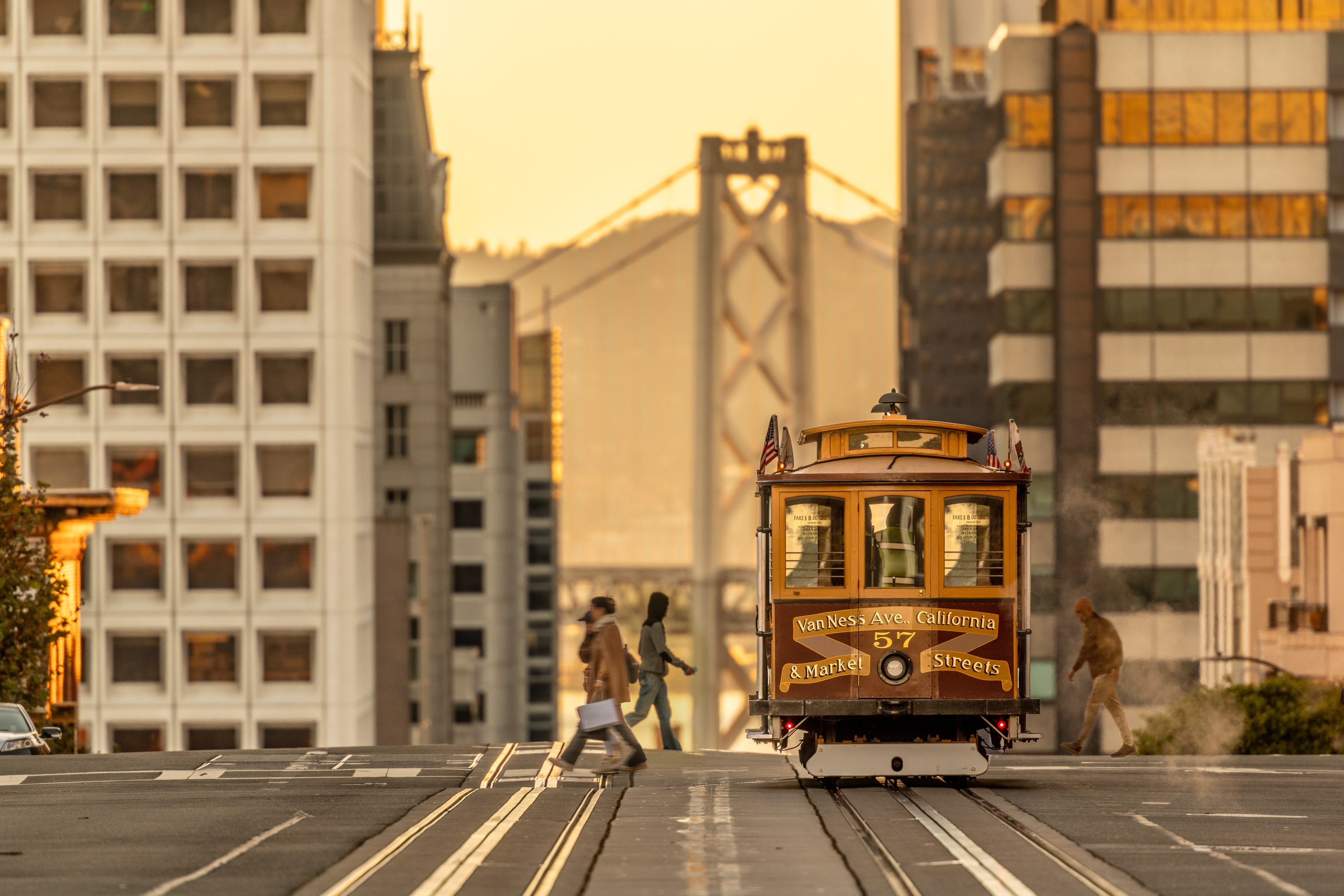 A San Francisco cable car travels down a city street with people crossing and a bridge visible in the background