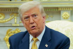 A person in a suit and tie sits in an ornate room, gesturing during a conversation. A fireplace and a bust are visible in the background