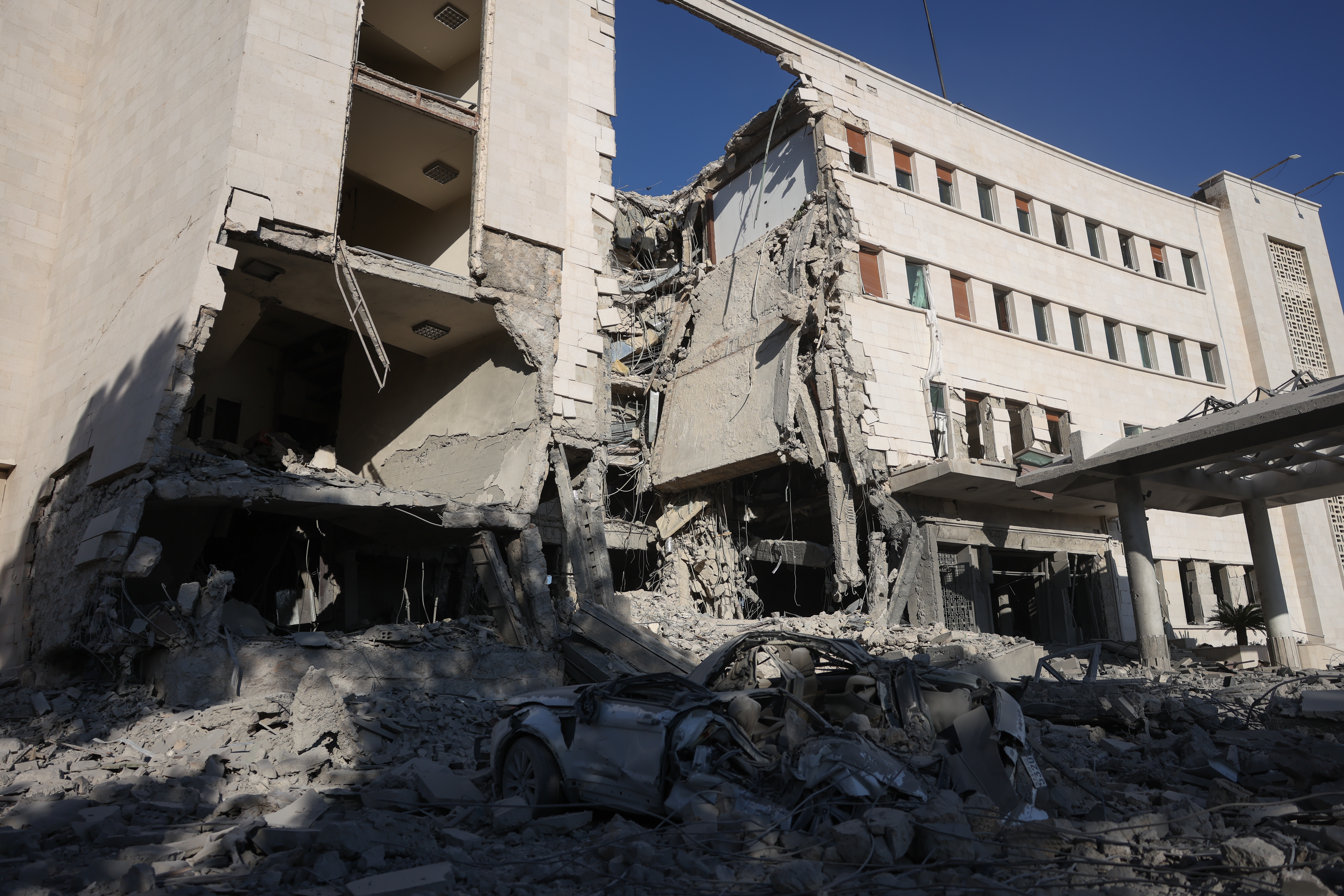 Partially collapsed building with debris scattered, showing extensive damage. A damaged car is in the foreground. Scene suggests recent destruction