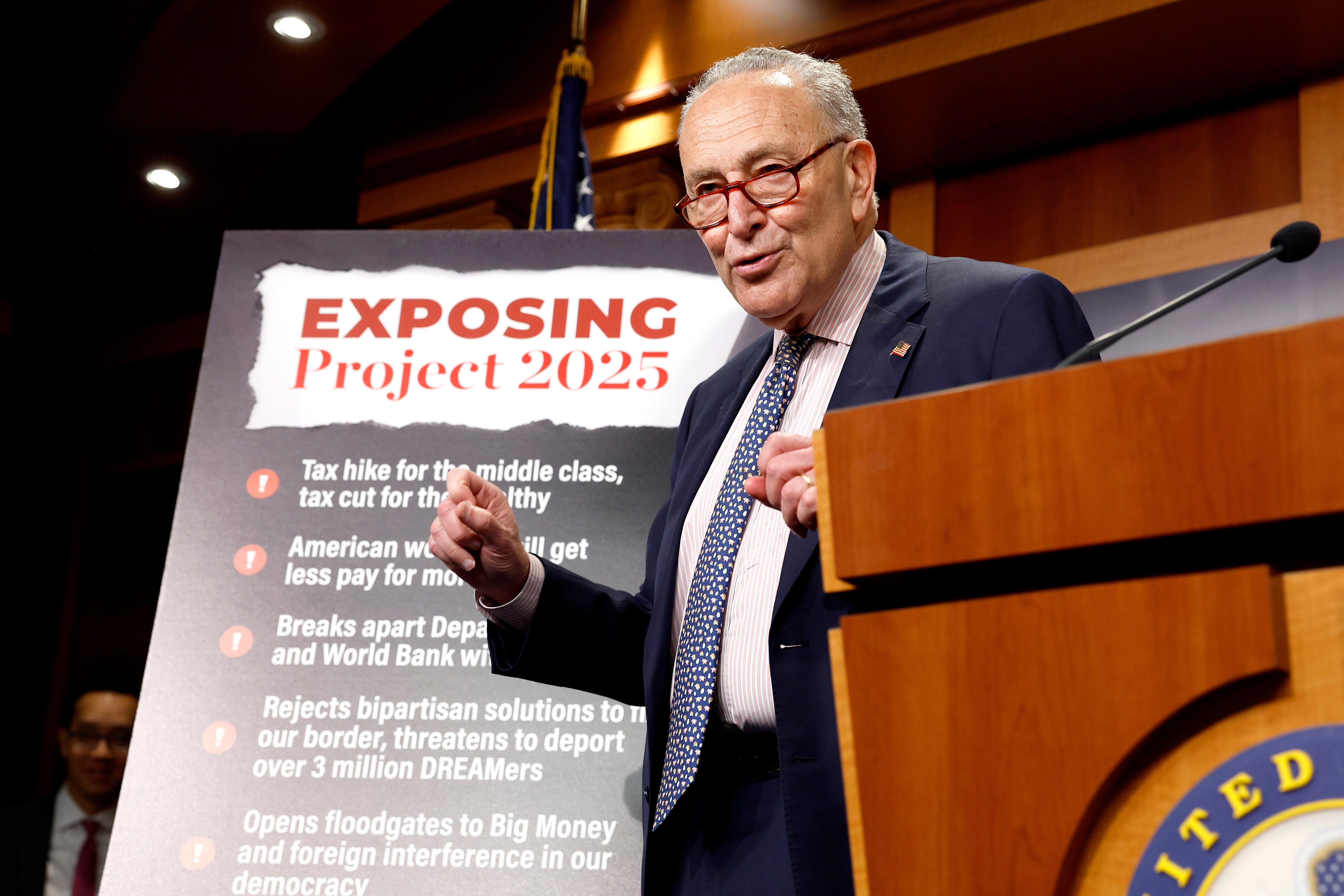 A man in a suit speaks at a podium with a sign titled &quot;Exposing Project 2025,&quot; listing various political critiques