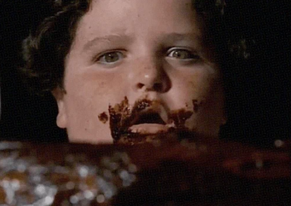 A child with chocolate smeared on their face looks up, seemingly caught mid-bite. Appears to be an indulgent moment involving food