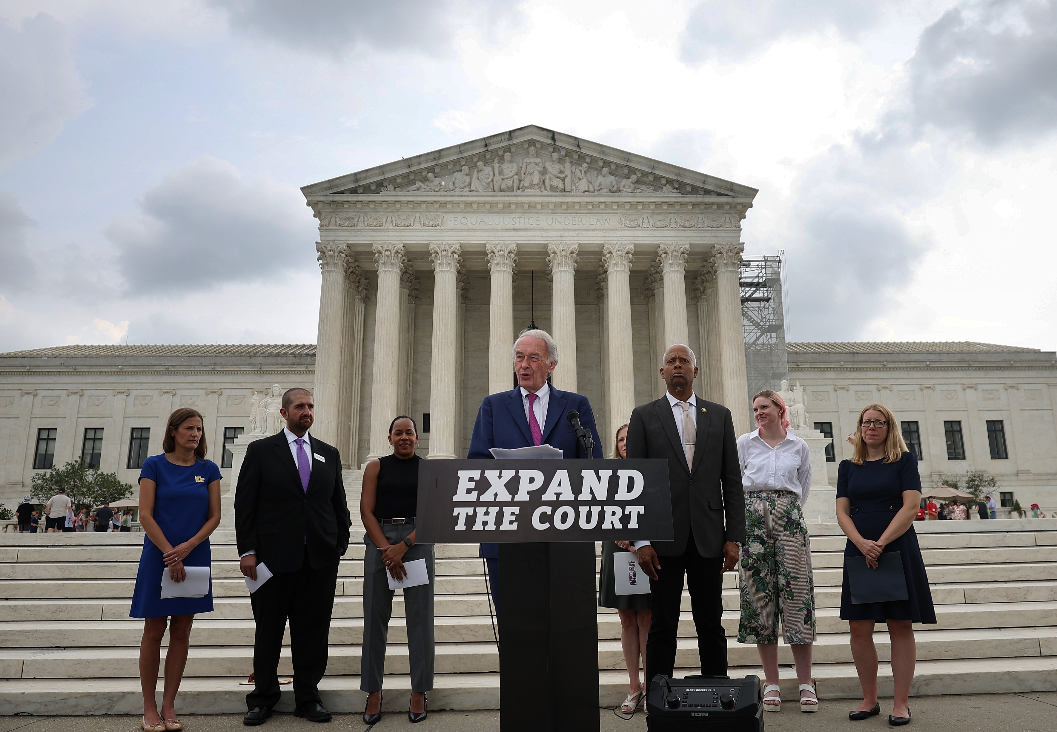 People standing on steps in front of a courthouse, holding an &quot;Expand the Court&quot; sign, dressed in formal attire