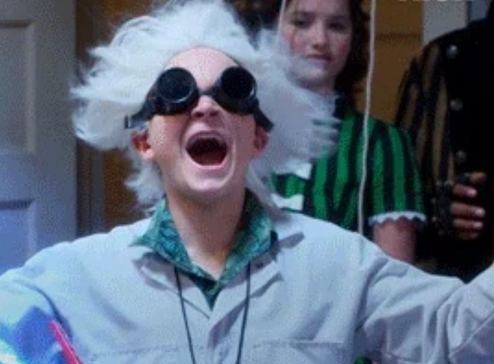 Person in a white lab coat with wild hair and goggles, smiling widely. Another person in a vintage-style dress is in the background