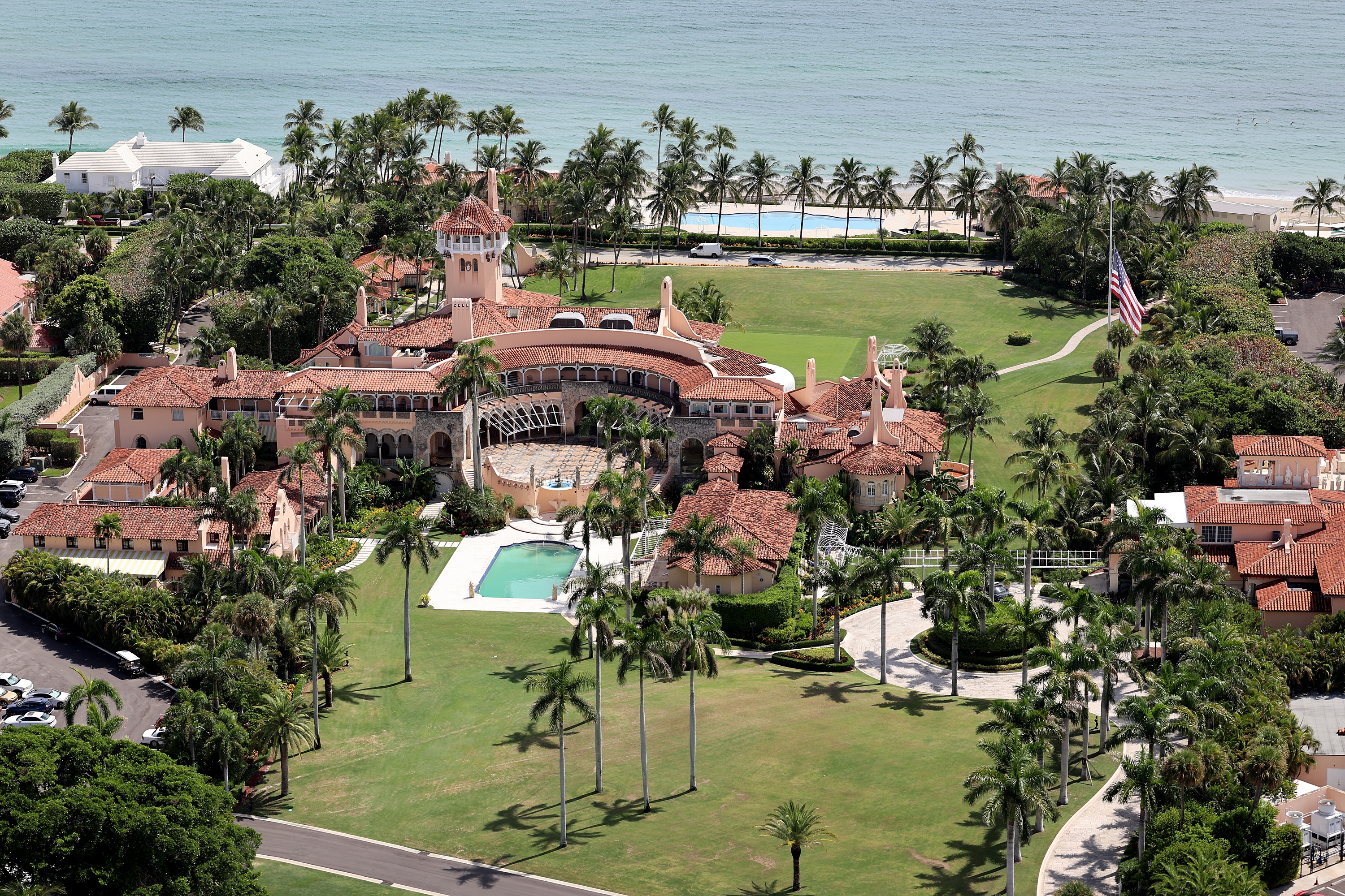 Aerial view of a large estate with red-tiled roofs, multiple buildings, and a central pool, surrounded by palm trees near the ocean
