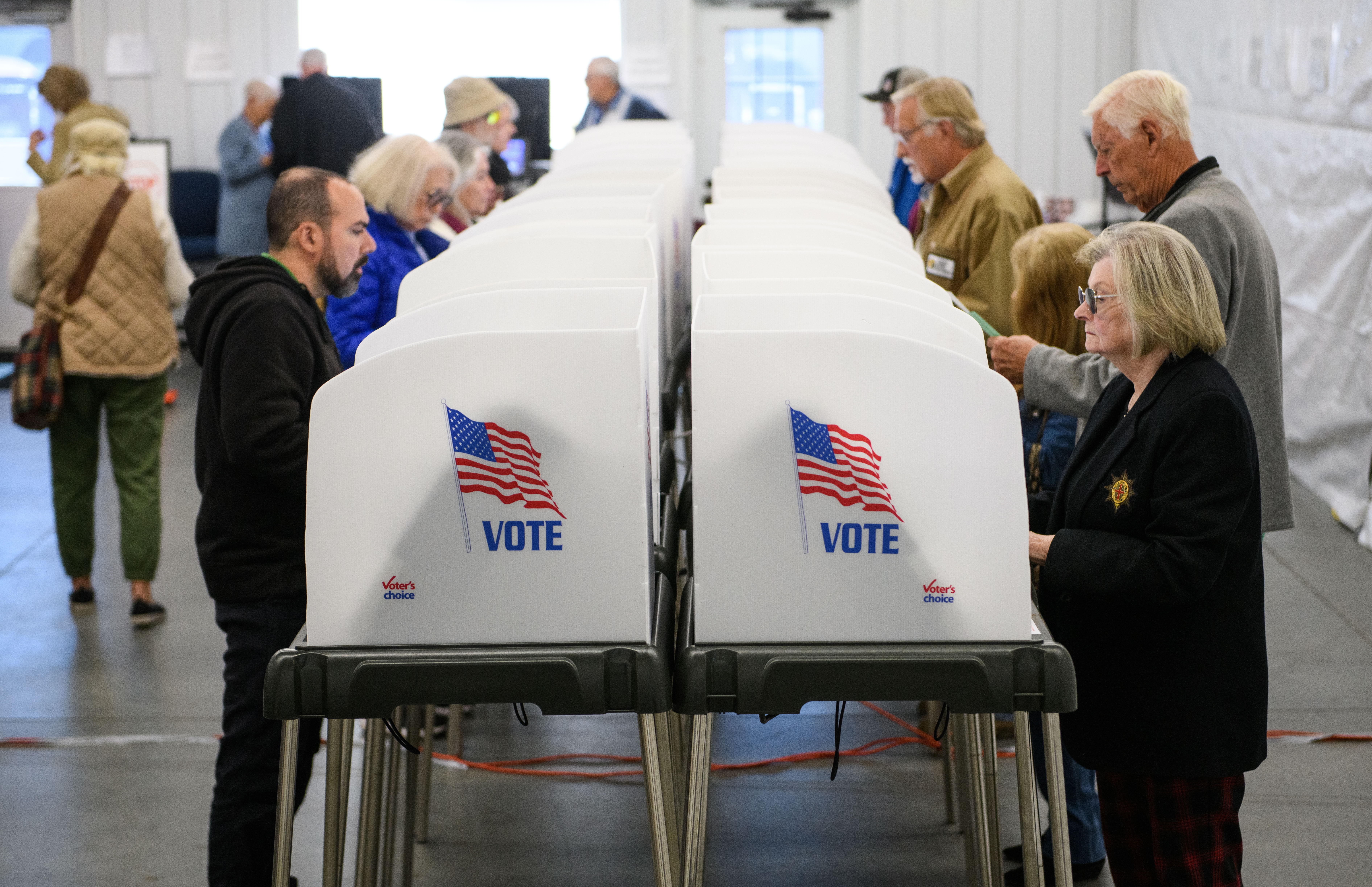 People voting at polling stations in a room, focusing on casting their votes. American flag and &quot;Vote&quot; visible on booths
