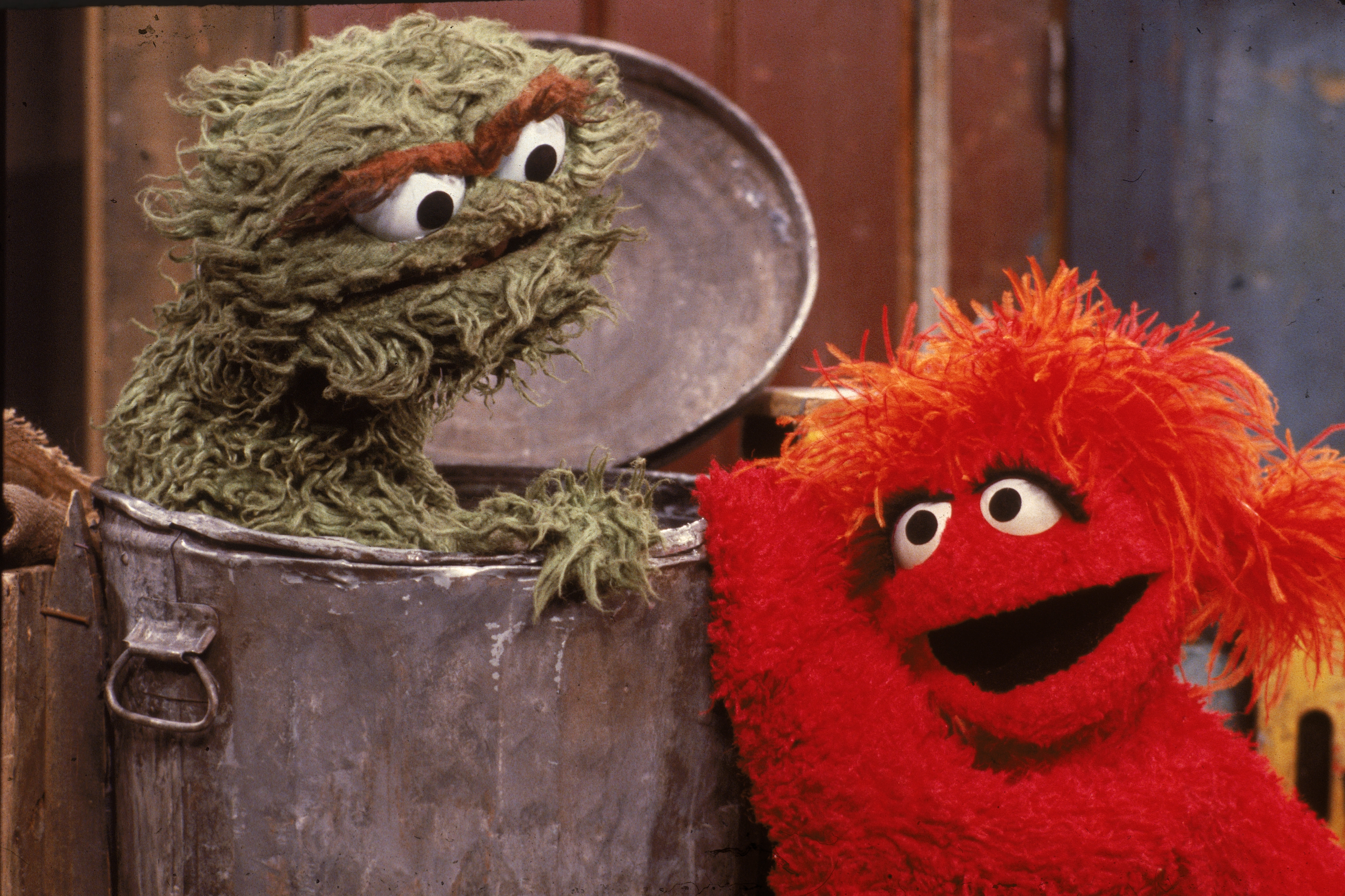 A green puppet in a trash can interacts with a red furry puppet, both showing expressive faces, set against a wooden background