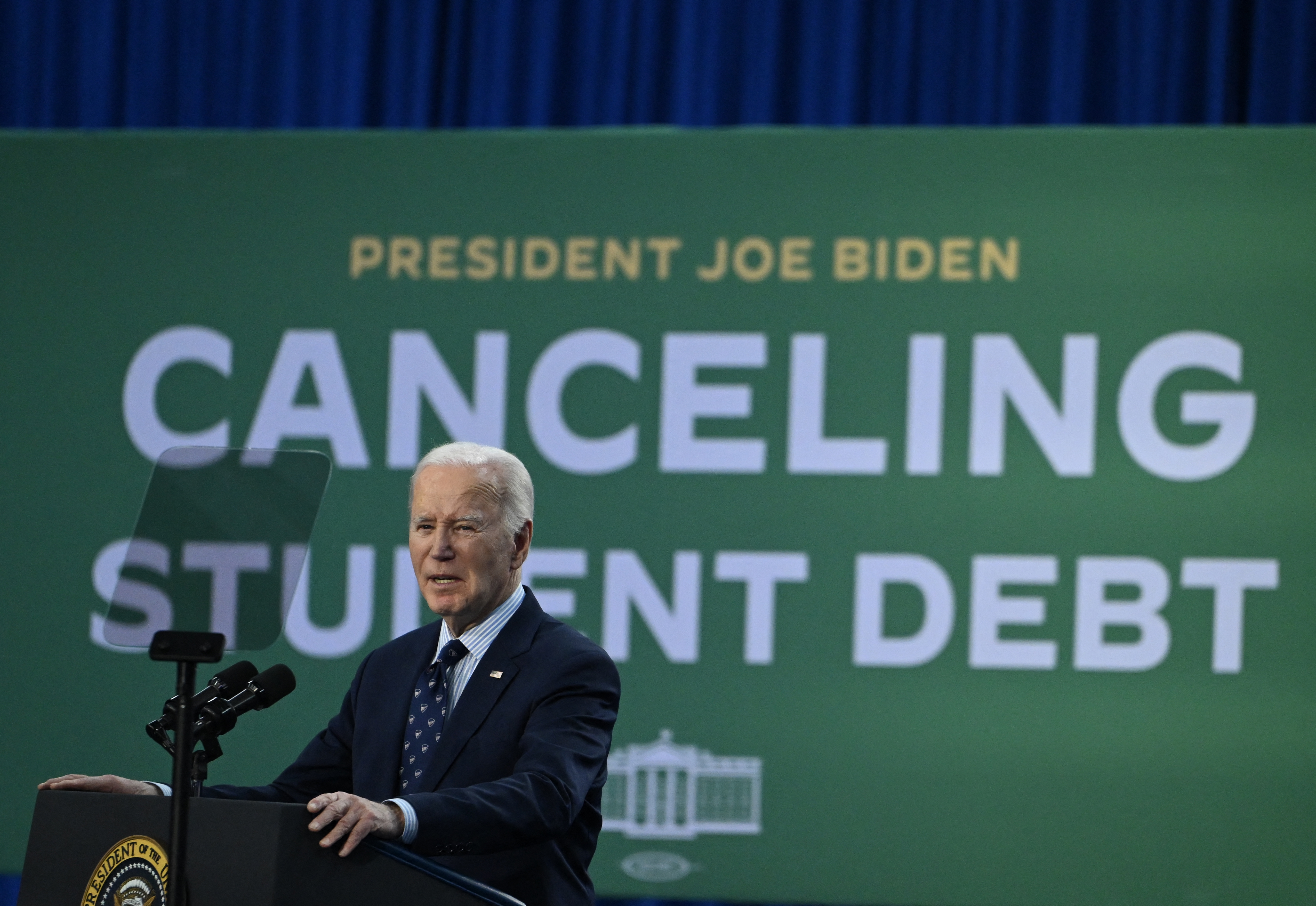 President Joe Biden speaks at a podium in front of a large sign reading &quot;Canceling Student Debt.&quot;