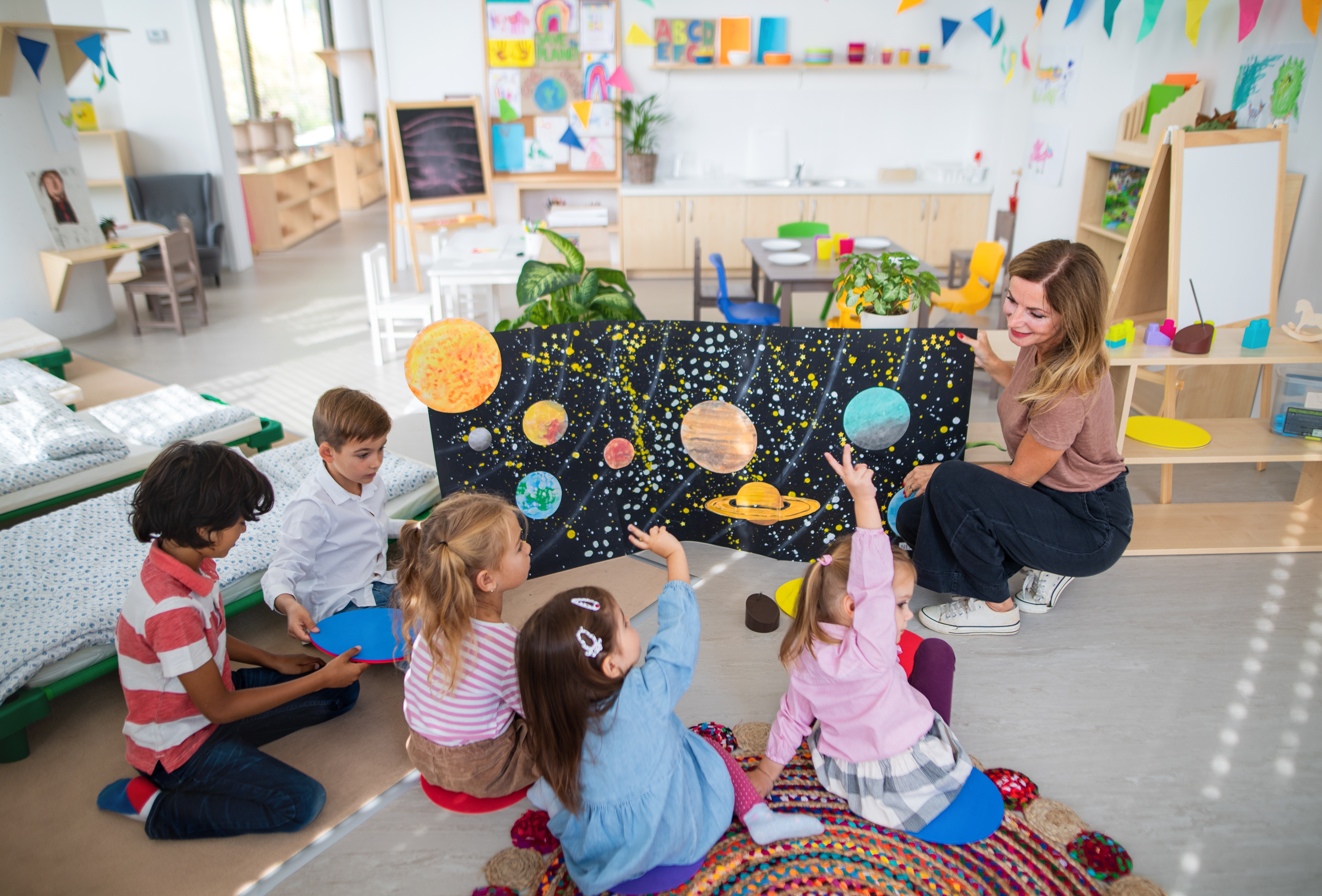 Children and a teacher discuss a solar system model in a classroom, engaging with planets on a colorful poster