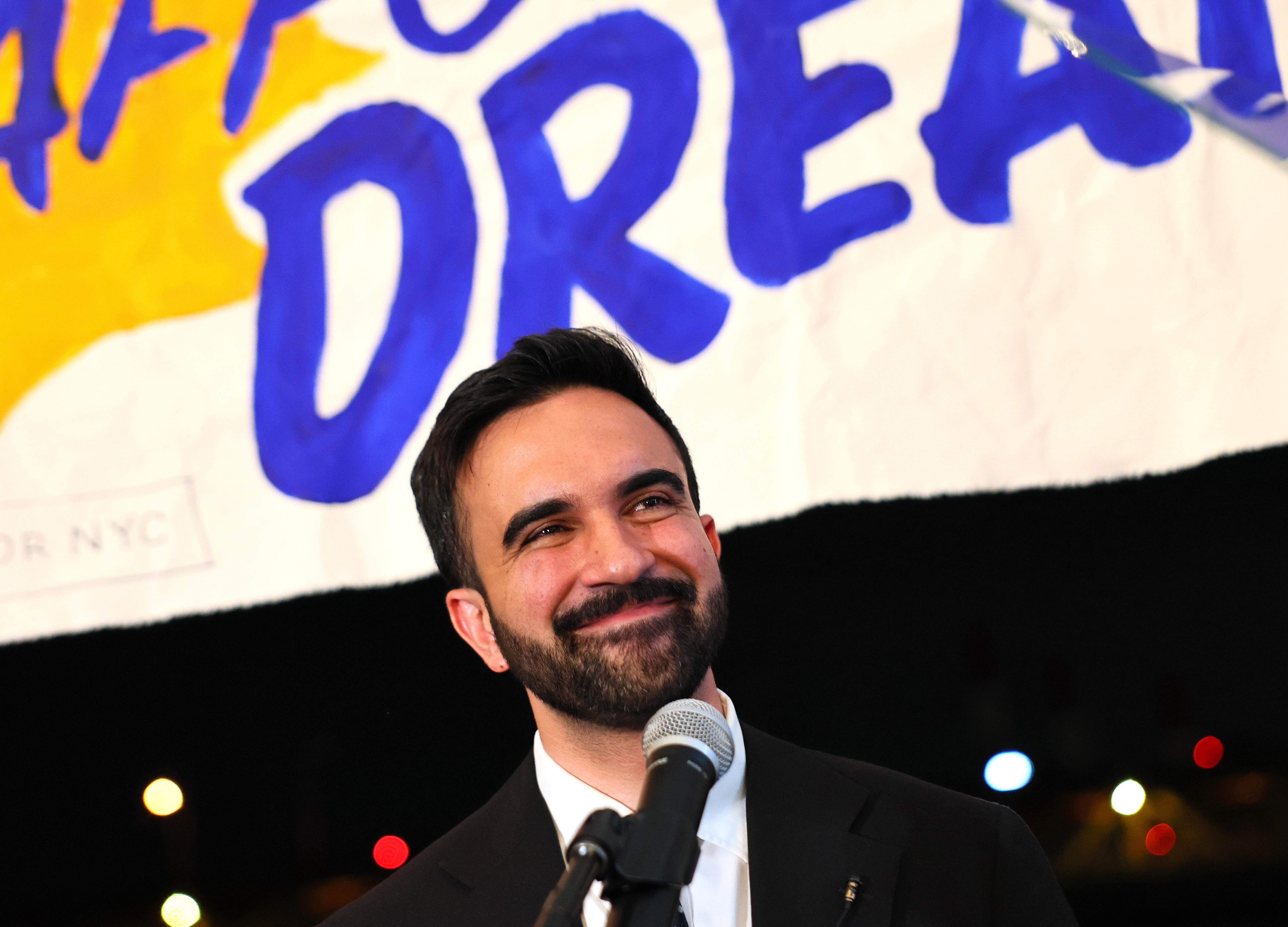 Man smiling at microphone, wearing a dark suit, with a sign in the background reading &quot;Dream.&quot;