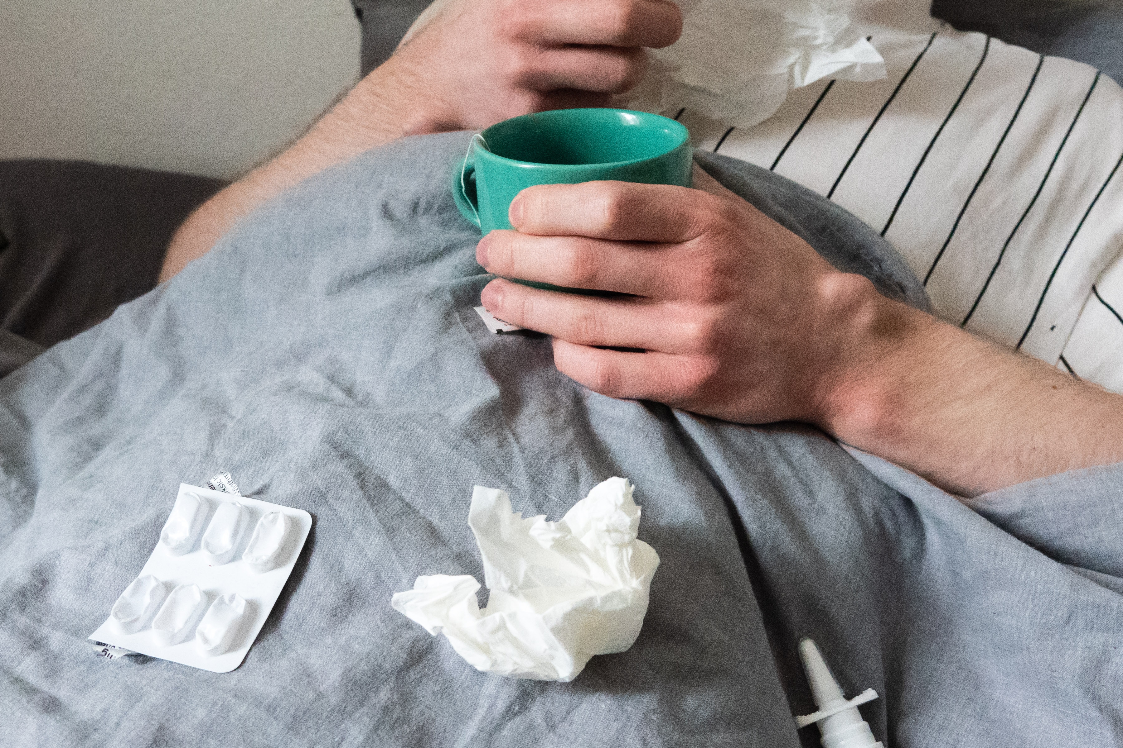 Person in bed holding a mug, surrounded by tissues and medication blister pack, suggesting they might be unwell