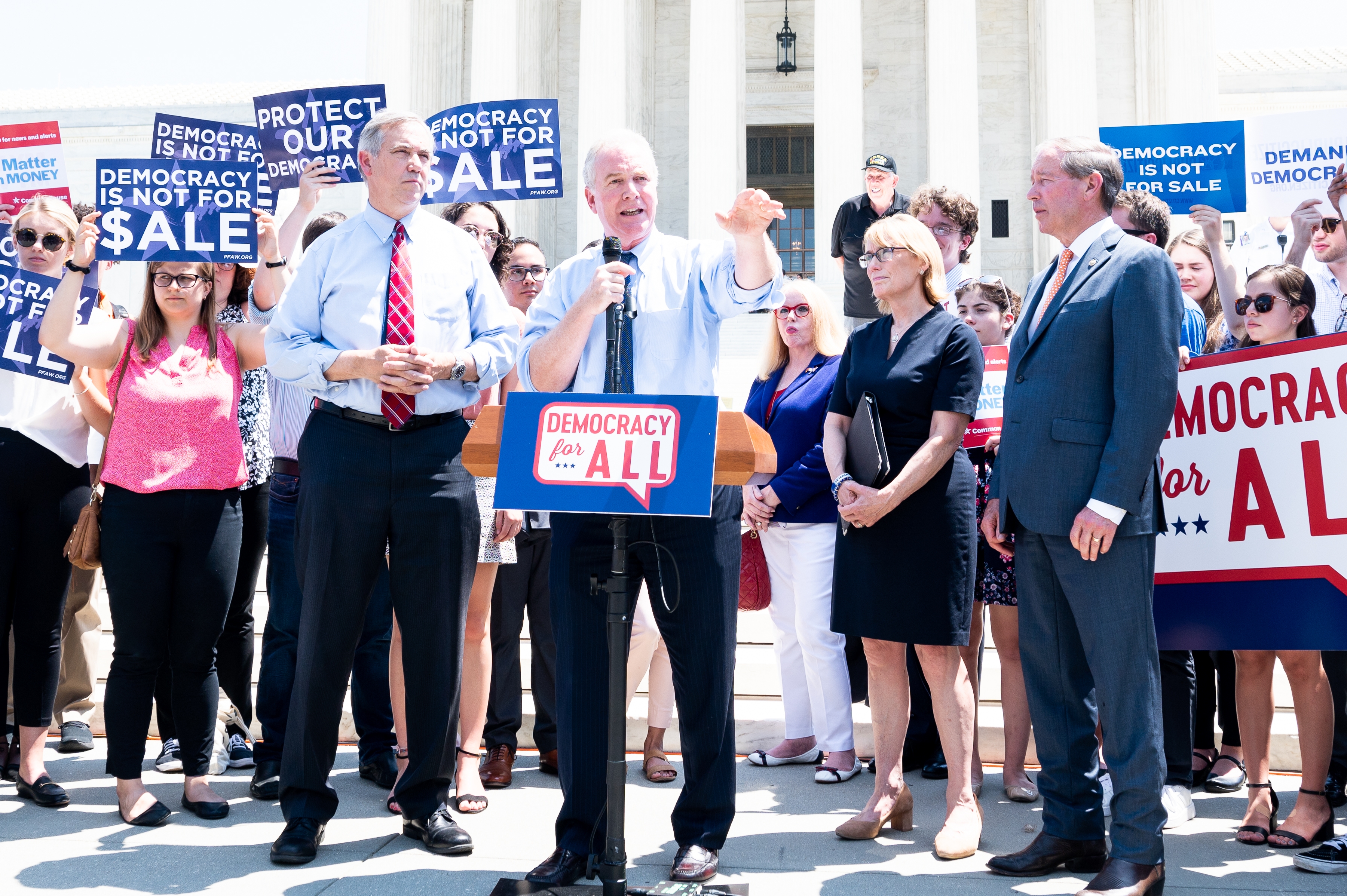 Group of people at a protest holding signs that say &quot;Democracy is Not for Sale&quot; and &quot;Democracy for All&quot; outside a government building