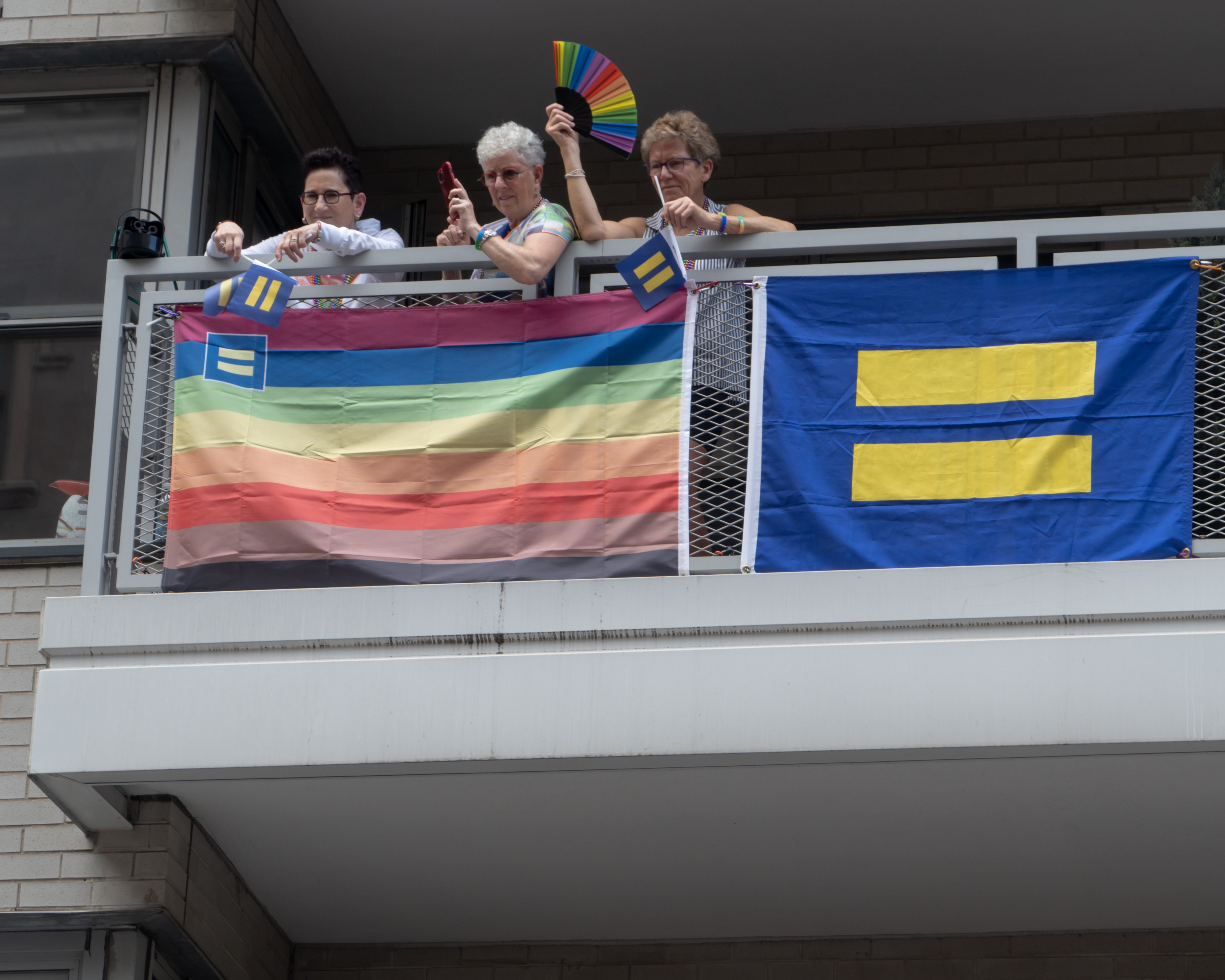 Three people stand on a balcony with rainbow and equality flags, holding rainbow fans, participating in a LGBTQ+ pride event