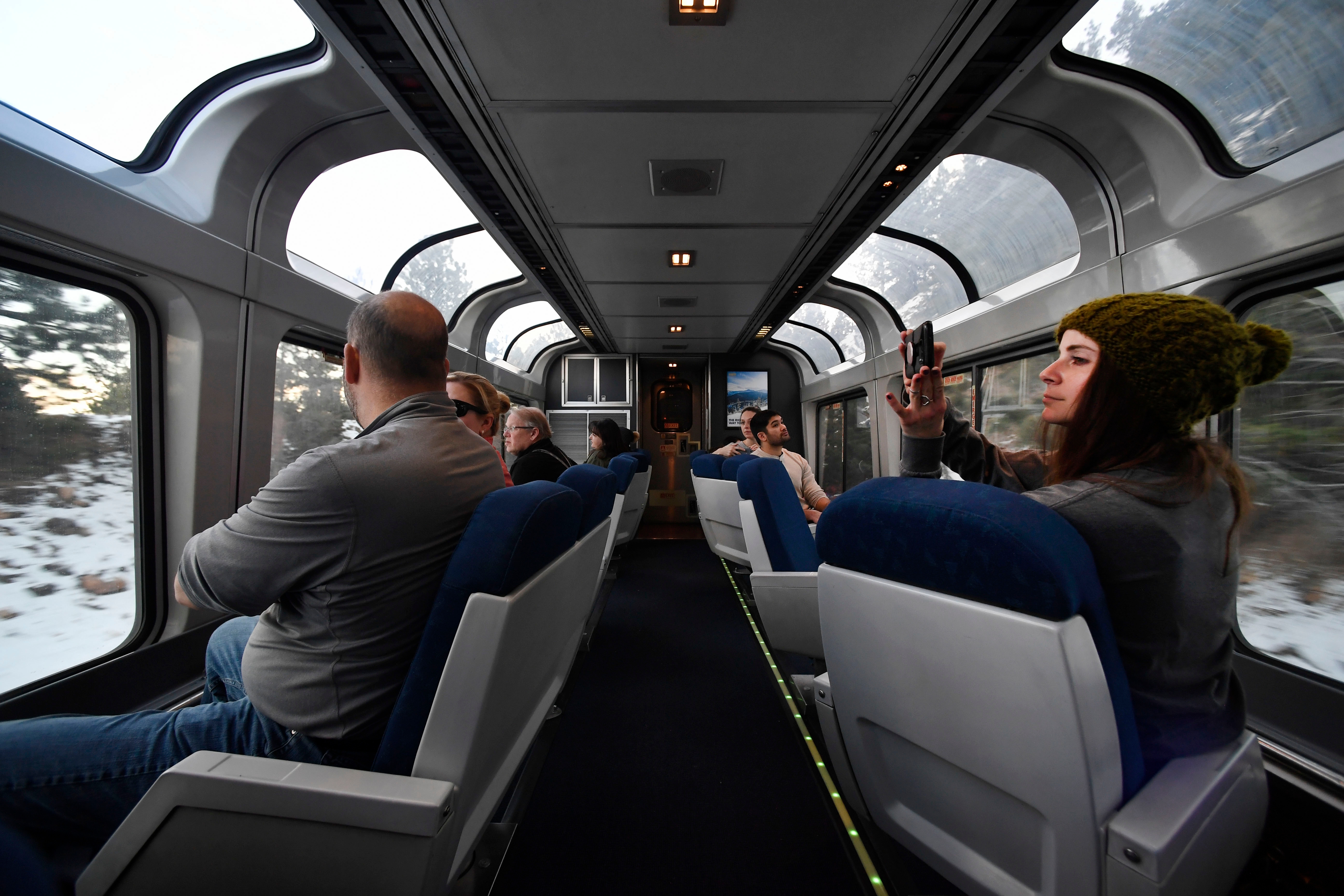 Passengers sit and look out large windows in a train observation car, with one person taking a photo of the scenic view outside