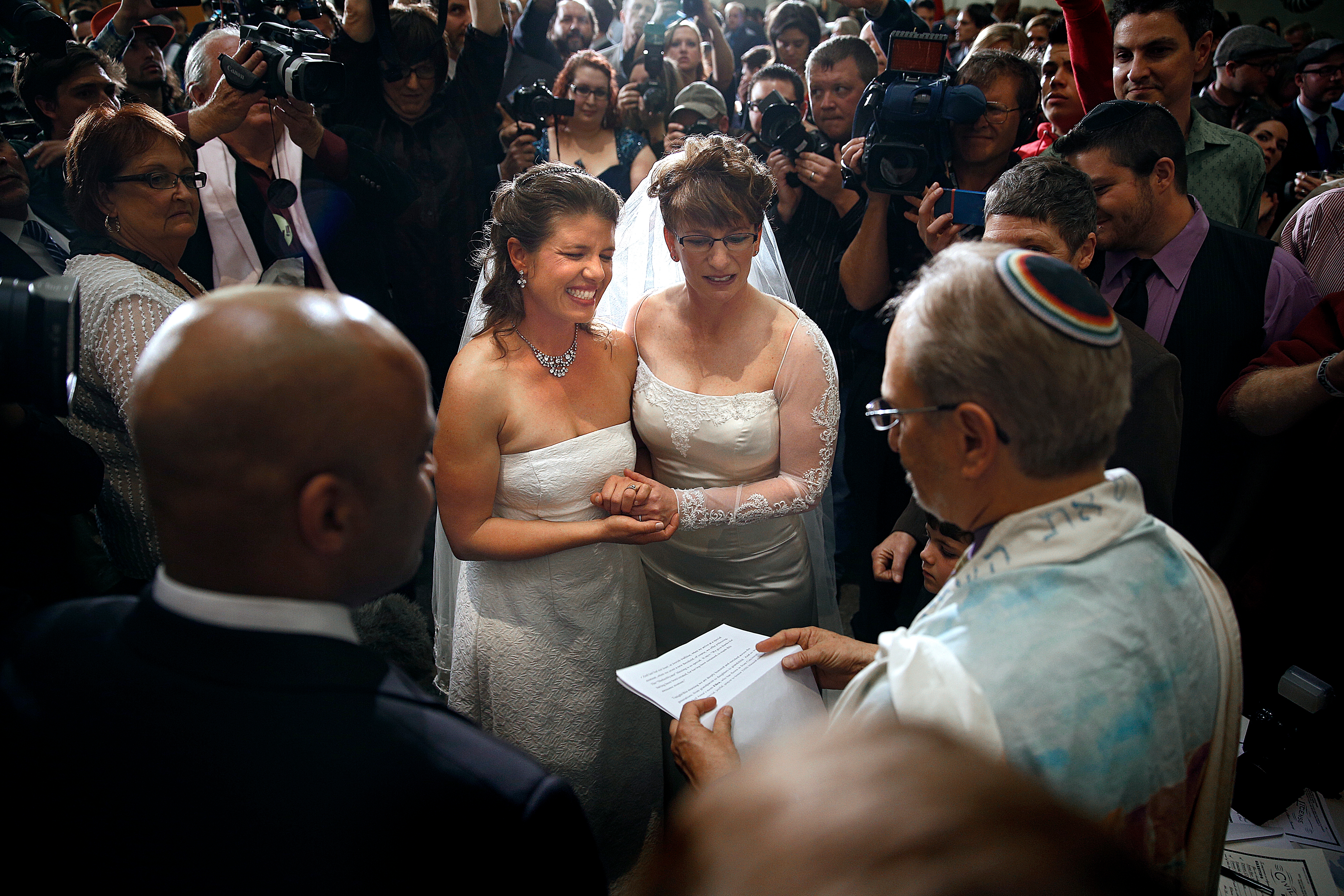 Two brides in lace dresses hold hands, smiling during a wedding ceremony surrounded by guests and photographers