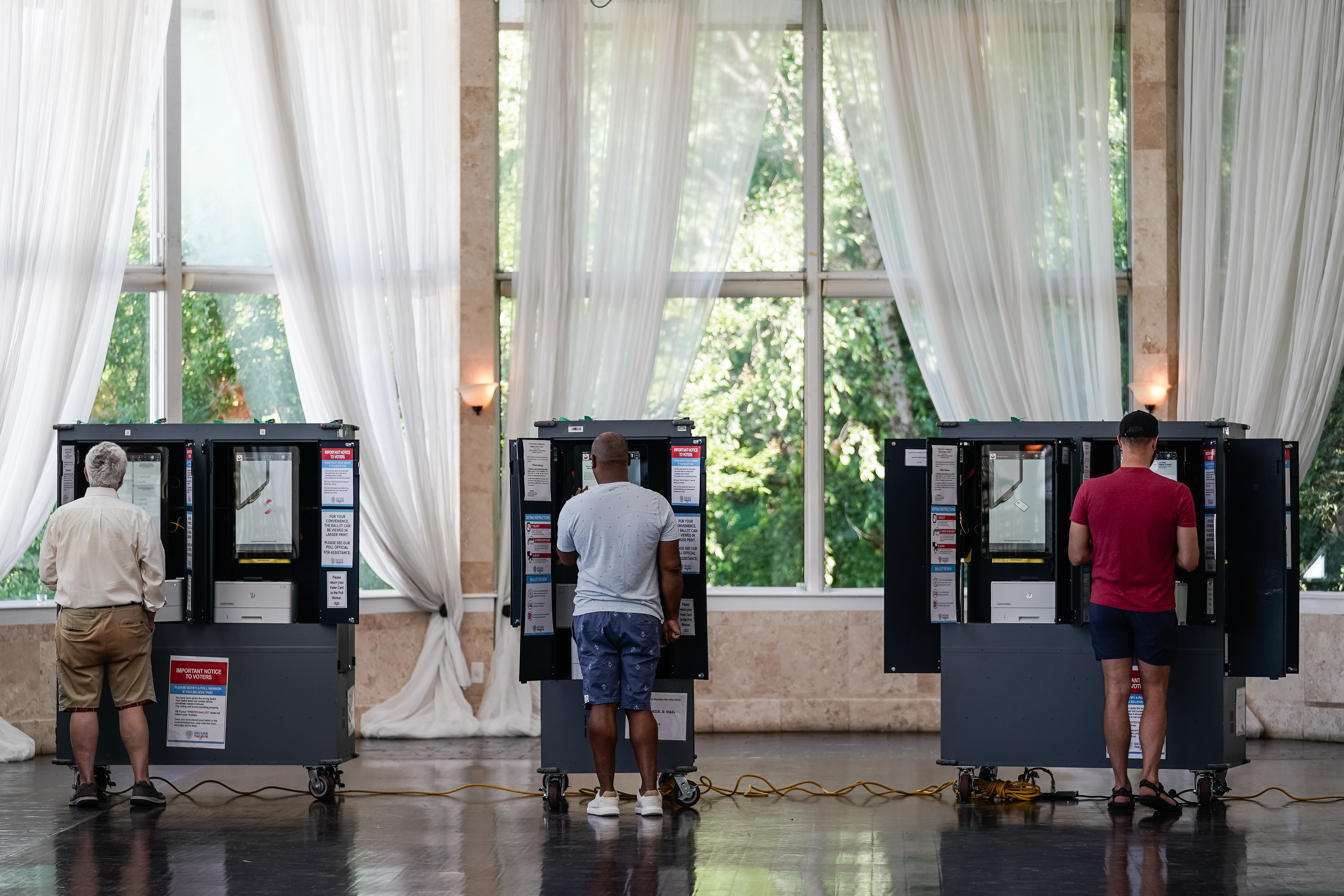 Three people vote at individual booths in a well-lit room with large windows and sheer curtains