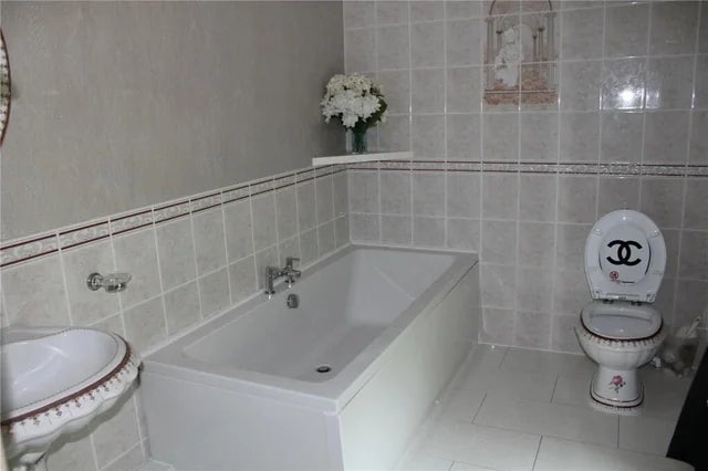 Bathroom with achromatic  tiled walls, featuring a bathtub, sink, and toilet. Toilet spot   has a Chanel logo. Flowers are placed connected  a ledge
