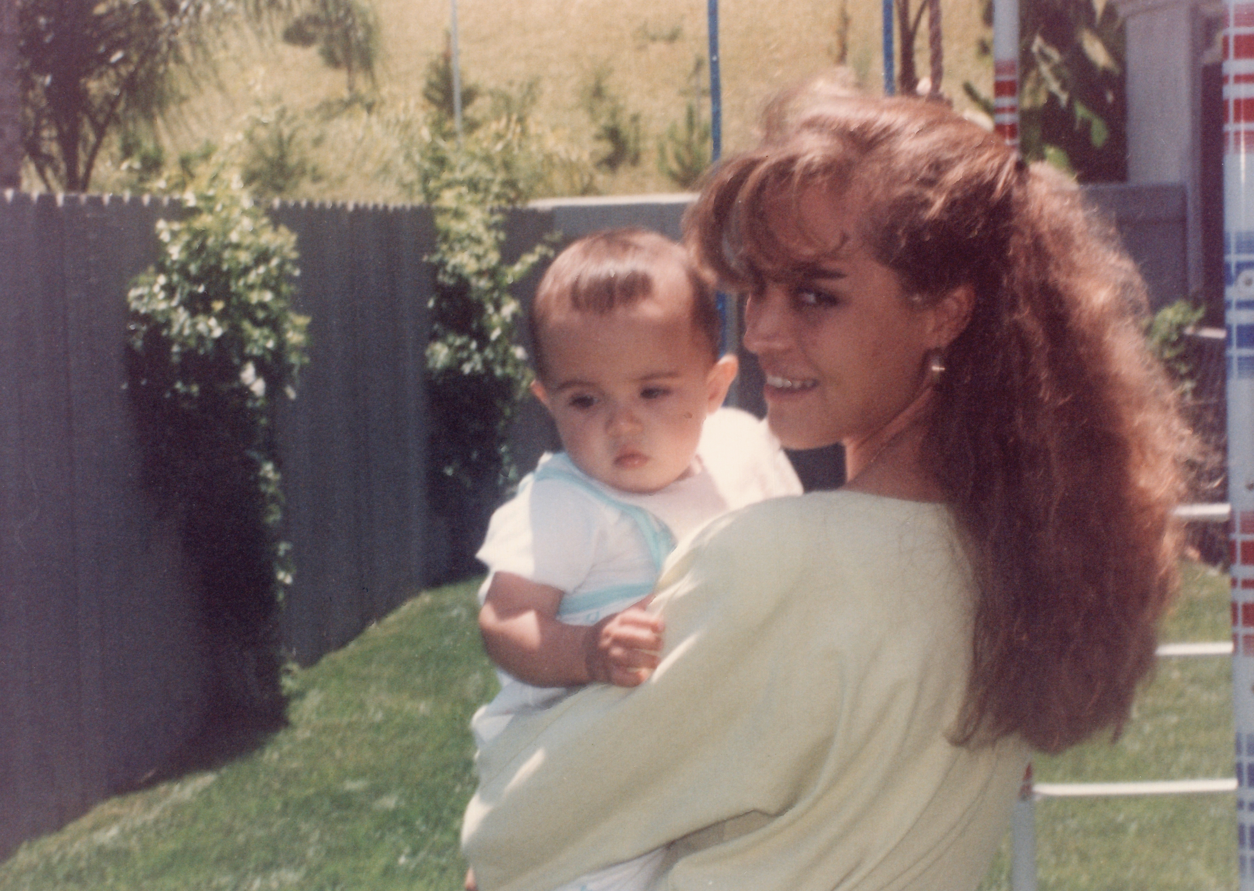 En kvinne med langt hår holder en baby i en hage, begge smiler til hverandre