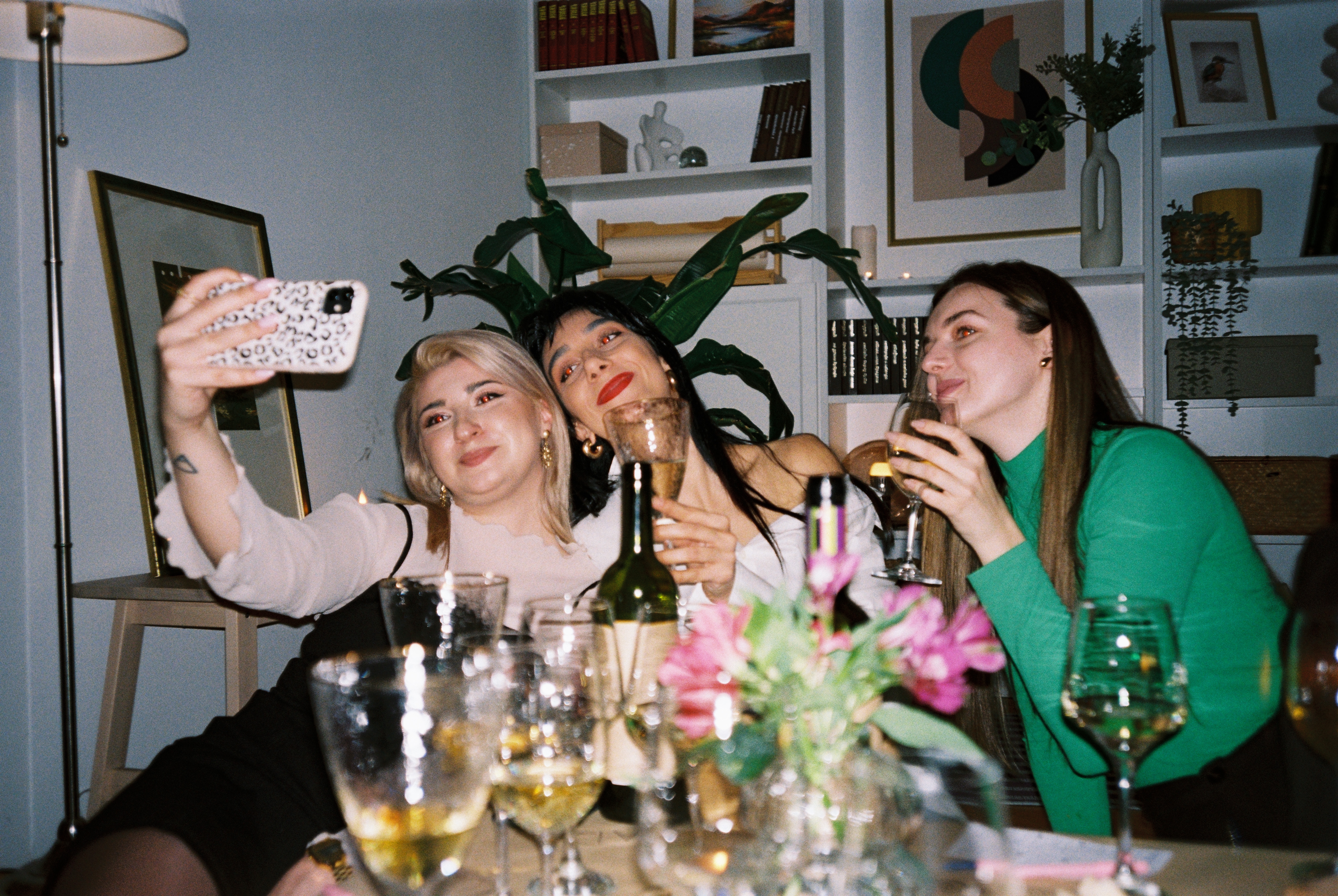Tre kvinner som sitter ved et bord, tar en selfie mens du holder vinglass, med blomster og dekorasjoner i bakgrunnen
