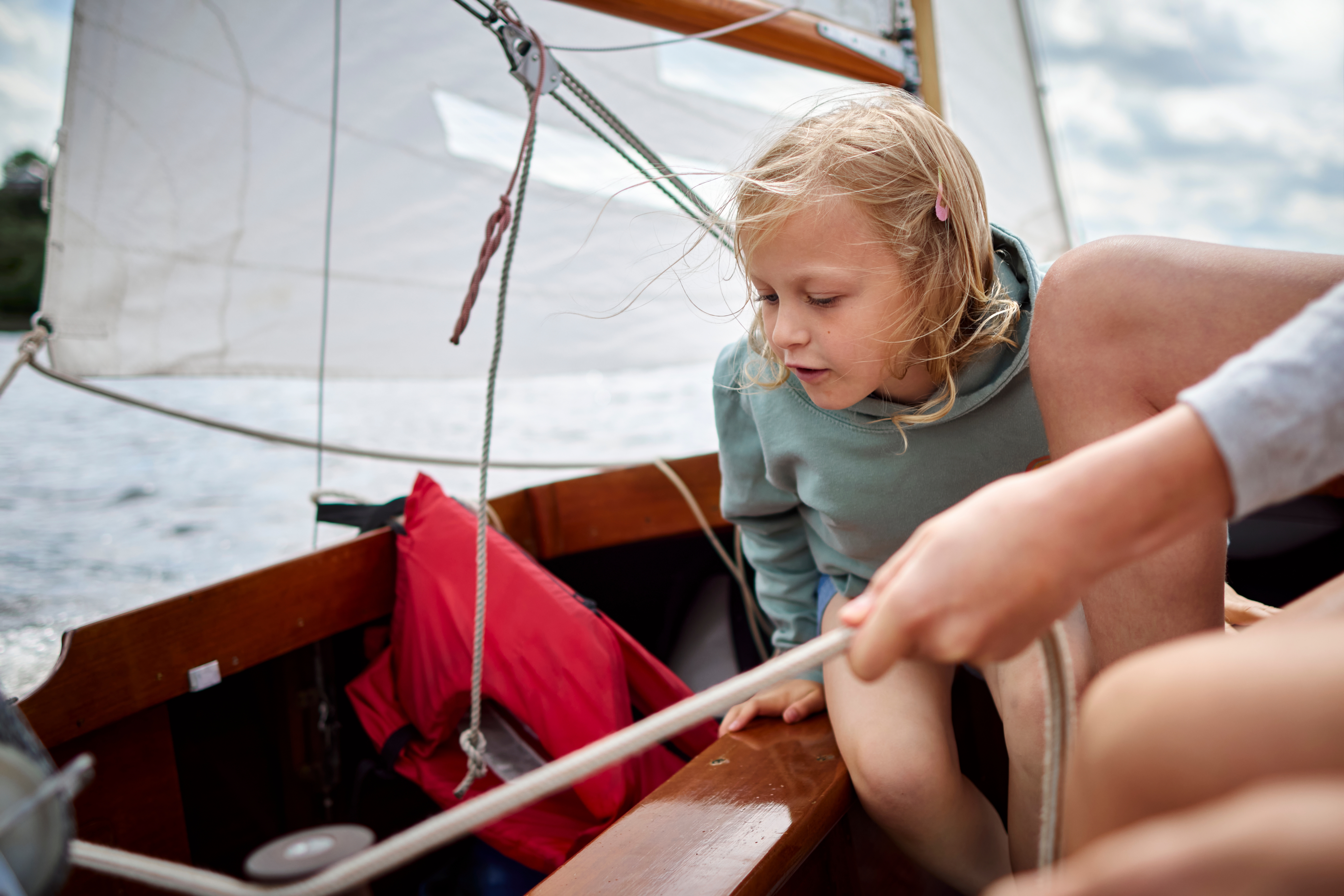Barn på en seilbåt, fokusert på vannet, mens en annen person administrerer tauet, og antyder en delt seilopplevelse